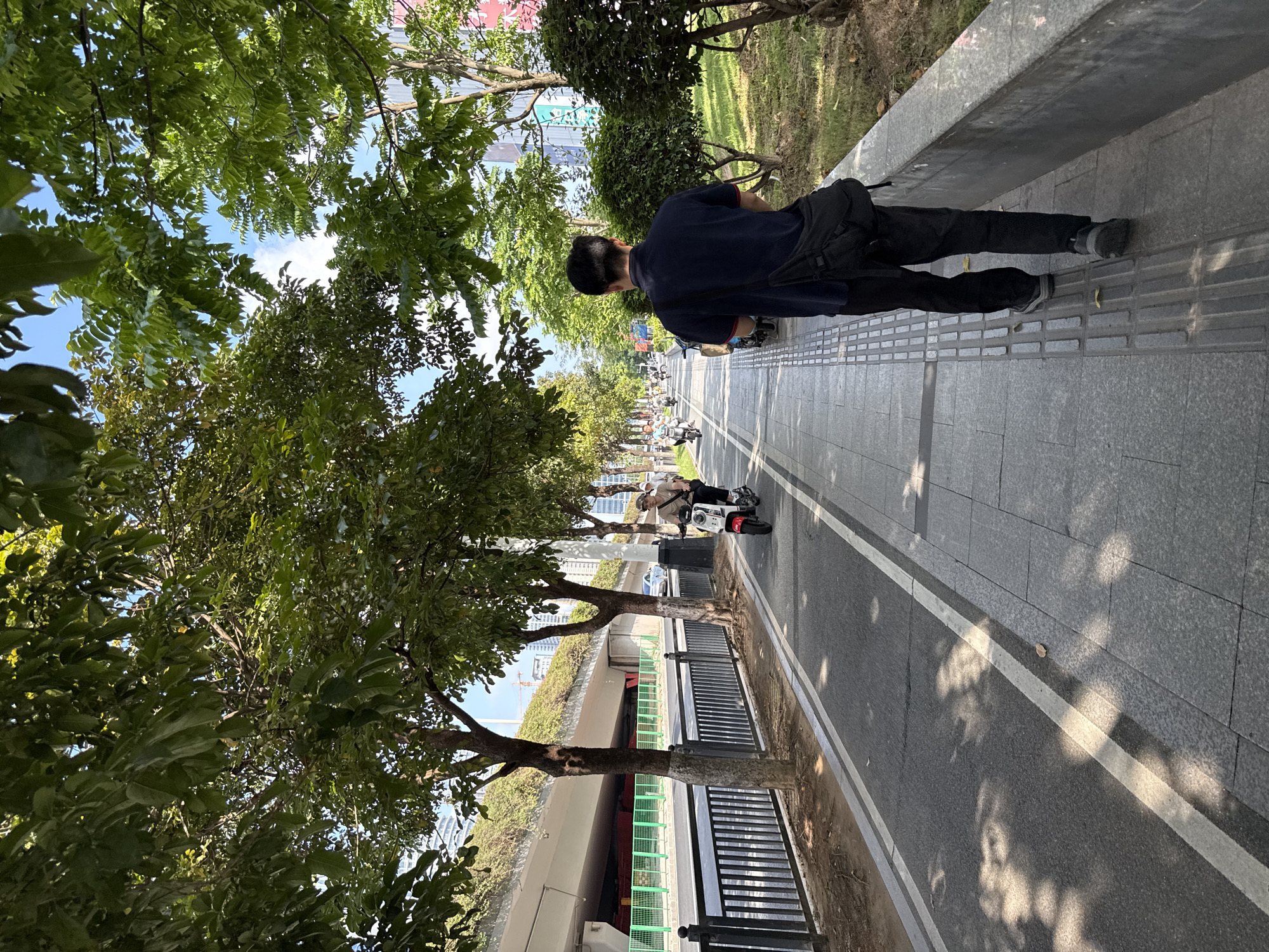 In Tianhe District, China, a pedestrian walks on a sidewalk next to a bicycle lane and a road. The man, viewed from behind, wears a dark blue polo shirt and black pants, carrying a black messenger bag over his right shoulder. He walks towards the right side of the frame. A person on a white electric scooter, wearing a light-colored top and a cap, rides in the bicycle lane ahead of him. The road to the left features vehicles, and a large elevated concrete structure with greenery on top is visible in the background, suggesting an urban highway or overpass. Trees with lush green foliage line the sidewalk, casting dappled shadows on the pavement. The sky is clear and blue.
