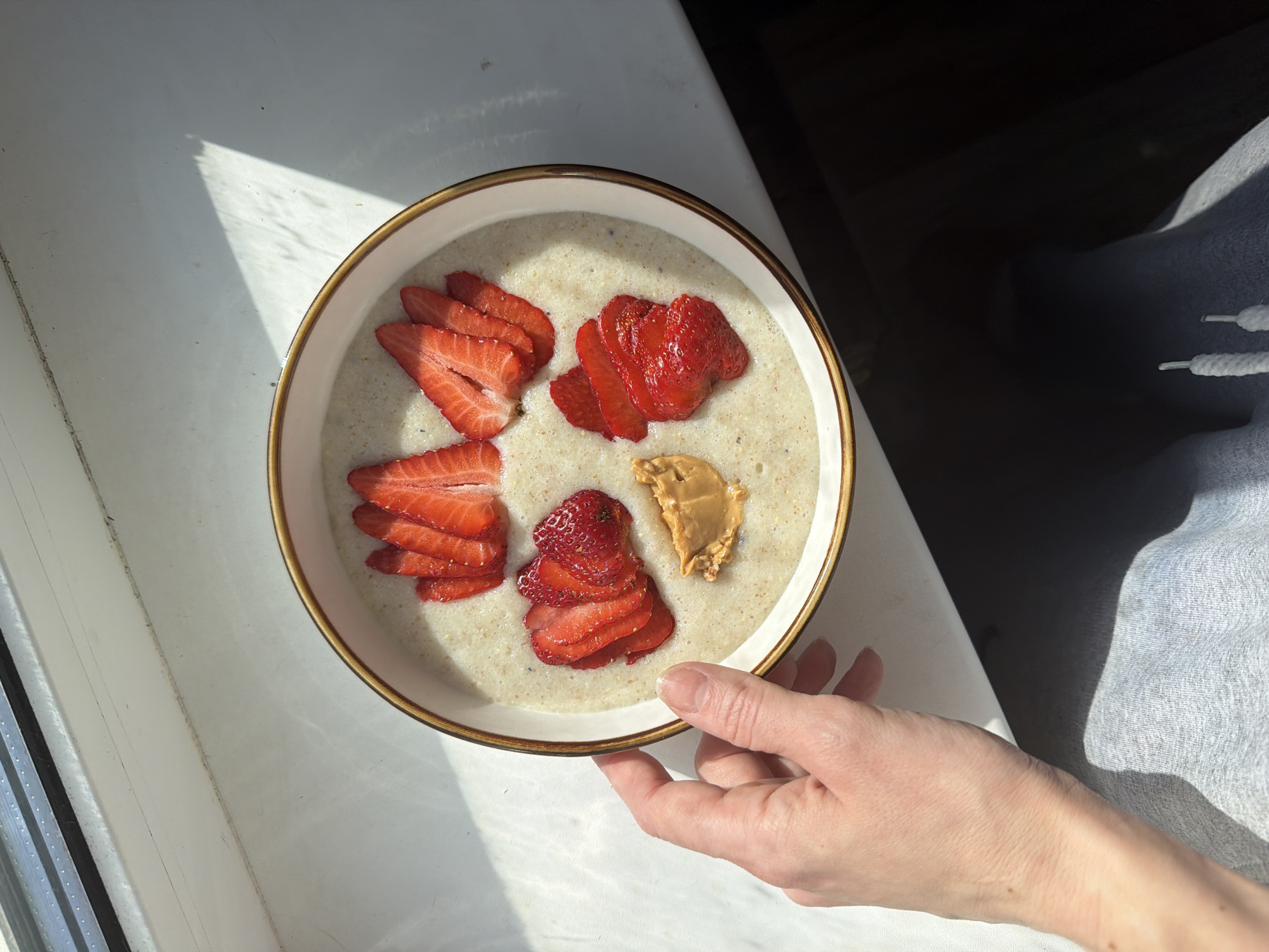 A high-angle, full-shot depicts a bowl of porridge with strawberries and peanut butter held by a person's hand, likely photographed in Perm, Russia. The bowl, primarily white with a gold rim, is filled with a light-beige porridge. Sliced strawberries are arranged in four separate piles around the perimeter, and a dollop of peanut butter sits near the center of the bowl. The person's hand, with fair skin, is holding the bottom of the bowl. The bowl is placed on a white surface, likely a table or countertop, near a window with bright sunlight casting shadows across the scene. Part of the person's arm and light-gray sweatpants are visible on the right side of the image.