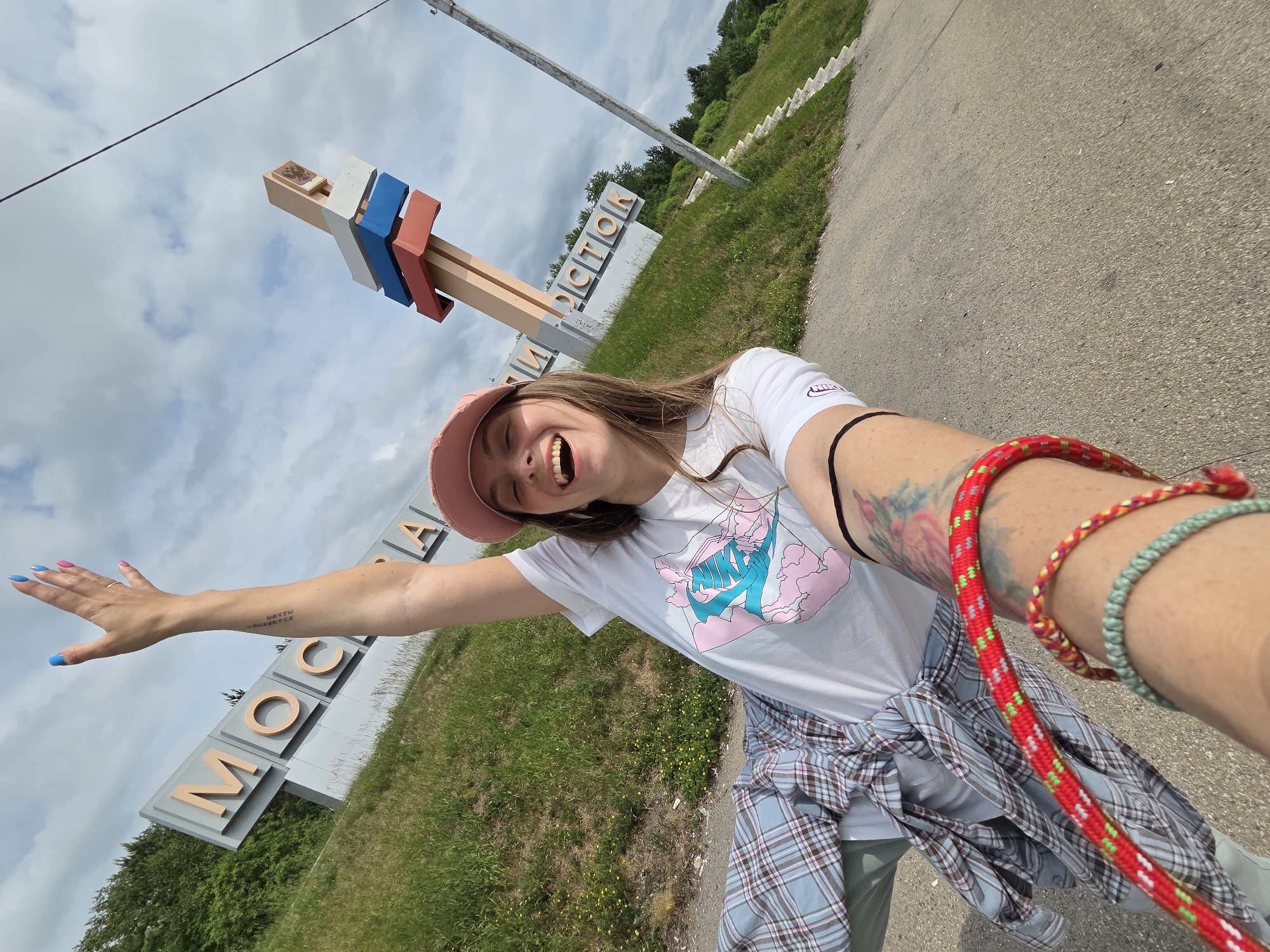 A young woman is taking a selfie on a cloudy day in Nikolaevka, Russia, in front of a large sign that says "MOSCOW". She is wearing a pink baseball cap, a white t-shirt with a graphic design, and a plaid shirt tied around her waist. Her right arm is raised in the air, and she is smiling broadly. She has tattoos on her arm and is wearing bracelets. The sign is large and white with orange letters. Behind the sign, there are trees and a grassy hill. There is also a tall pole with colored flags on it. The ground in front of the sign is asphalt.