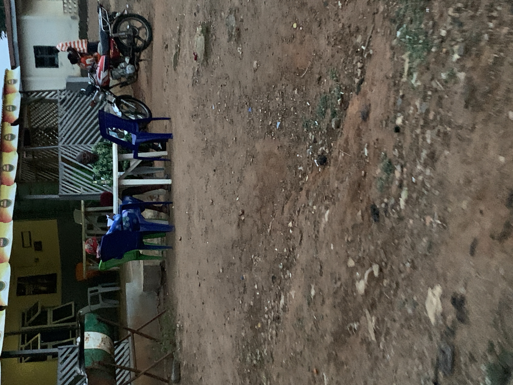 This image captures an outdoor scene in what appears to be a local establishment or gathering spot, likely at dusk or night, given the low light conditions. The ground is unpaved, dusty, and strewn with various pieces of litter and small debris, indicative of a well-used area.
Several subjects are visible. On the left side, two motorcycles are parked. One red and black motorcycle has a person standing or sitting near it, wearing a striped shirt. Another red motorcycle is parked slightly behind it. Closer to the foreground on the left, two blue plastic chairs are placed around a table, and another person is partially visible behind the table. Further to the left, within a covered area, several more blue and green plastic chairs are arranged around tables, with a green barrel in the foreground. Inside this structure, which seems to be a covered seating area or a part of a building, additional furniture and what might be wall decorations or displays are visible under artificial light. The word "HERO" is faintly visible on some stacked, light-colored objects on the far left, possibly crates or packaging.
The setting appears to be a semi-urban or rural area in Kabba, Nigeria, characterized by simple building structures with patterned fences or railings, and the unpaved ground. The overall atmosphere suggests a casual, communal outdoor space. No specific activity beyond people being present and seated is clearly discernible. The quality of the image is somewhat grainy, and it is rotated ninety degrees clockwise.