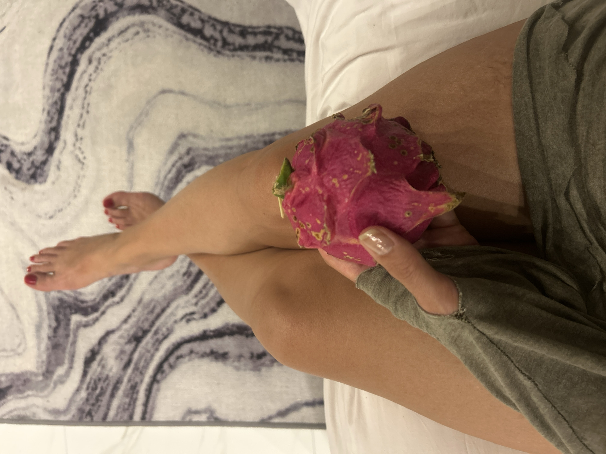 A woman is sitting on a white surface, possibly a bed, with her legs crossed. She is wearing a green shirt with a thumb hole and is holding a pink dragon fruit in her hand. Her toenails are painted red. The background includes a gray and white patterned rug. It is not possible to determine the location in Kediri, Indonesia, from the provided image.