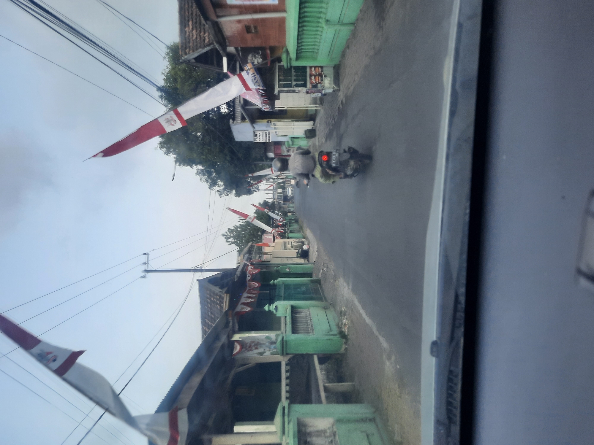 A medium shot captures a street scene in Banjarkemantren, Indonesia, with a focus on the right side of the road. The road itself is asphalted and appears to be a residential or commercial street. Along the right side, there are several small buildings with awnings and signs, suggesting shops or small businesses. One building has a sign that reads "PIJAT CAFE." Red and white Indonesian flags, characteristic of national celebrations, are prominently displayed, attached to poles and strung across the street. The flags are long and narrow, with the top portion being red and the bottom white, often bearing an emblem. A single motorcyclist, wearing a helmet, is riding away from the camera down the street on a dark-colored scooter. The background features trees and power lines, indicating an outdoor, daytime setting. The view appears to be from inside a vehicle, as the lower portion of the image shows the dashboard and windshield of a car. The lighting suggests it is early morning or late afternoon, with soft, diffused light. The overall atmosphere is quiet and depicts a typical street scene in a community that is likely celebrating a national holiday.