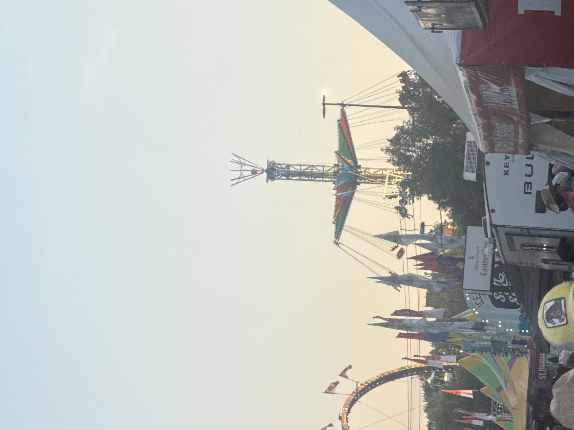 The image captures a scene at the Calgary Stampede, a well-known annual fair and rodeo held in Calgary, Alberta, Canada. The sky is a pale, hazy blue, suggesting either early morning or late afternoon light. In the background, a tall swing ride with colorful gondolas is in motion, with several people visible on it. To the left, a portion of a roller coaster with illuminated tracks can be seen curving upwards. Numerous flags, predominantly red, white, and blue, are planted in the foreground, some featuring Canadian maple leaves. Various tents and structures, typical of a fairground, are present, including a white trailer with the word "KEY" and "BUL" visible on its side, and a red banner with an emblem and text that appears to be "RACE". People are gathered in the lower part of the frame, some wearing hats, indicating a public event. Trees are visible behind the fairground structures, and a lamppost with a light on it stands near the swing ride. The overall atmosphere is that of a bustling outdoor festival.