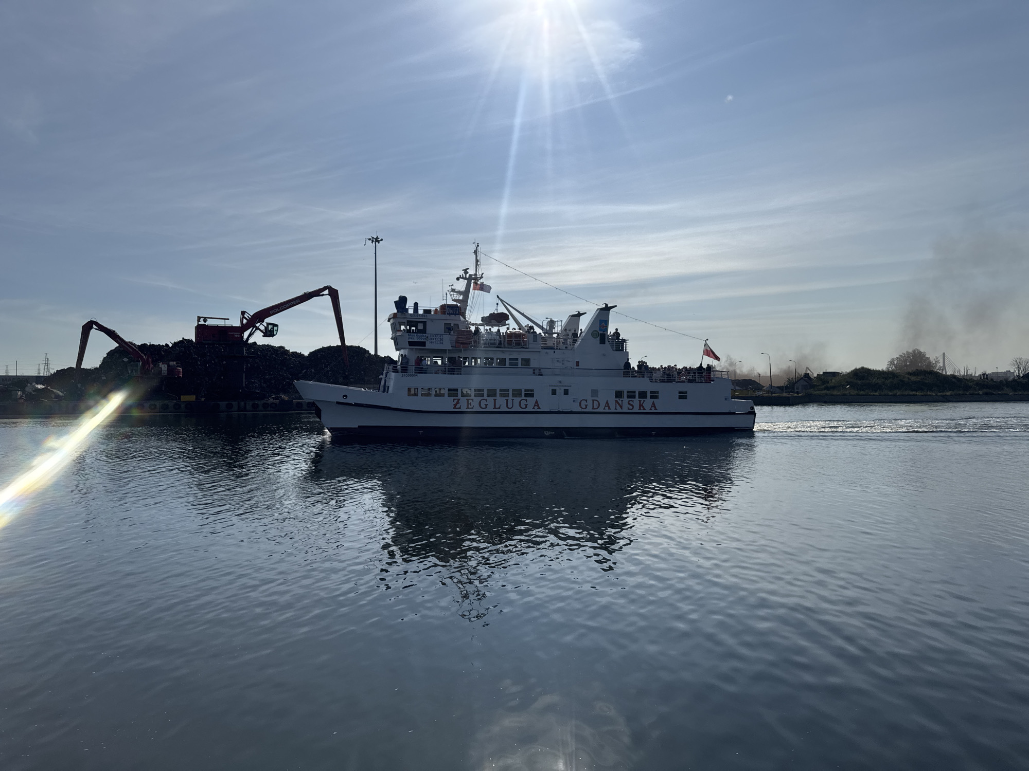 A ferry named "ŻEGLUGA GDAŃSKA" is sailing on a waterway in Gdańsk, Poland. The ferry is white with red lettering and has several decks with windows. People are visible on the upper deck. To the left of the ferry, there are industrial cranes unloading a large pile of dark material, possibly scrap metal, from a dock. The sky is bright with a sun flare. In the background, there are some buildings and trees, with a plume of smoke rising in the distance. The water is calm, with ripples and reflections.