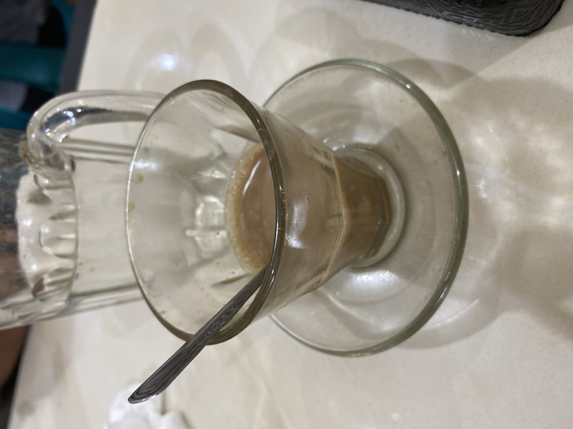 The image depicts a clear glass cup sitting on a clear glass saucer, with a spoon inside the cup. The cup contains a light brown liquid, possibly coffee with milk. A clear glass pitcher is partially visible to the left of the cup. The background appears to be a white or light-colored table. The location is Lhokseumawe, Indonesia.