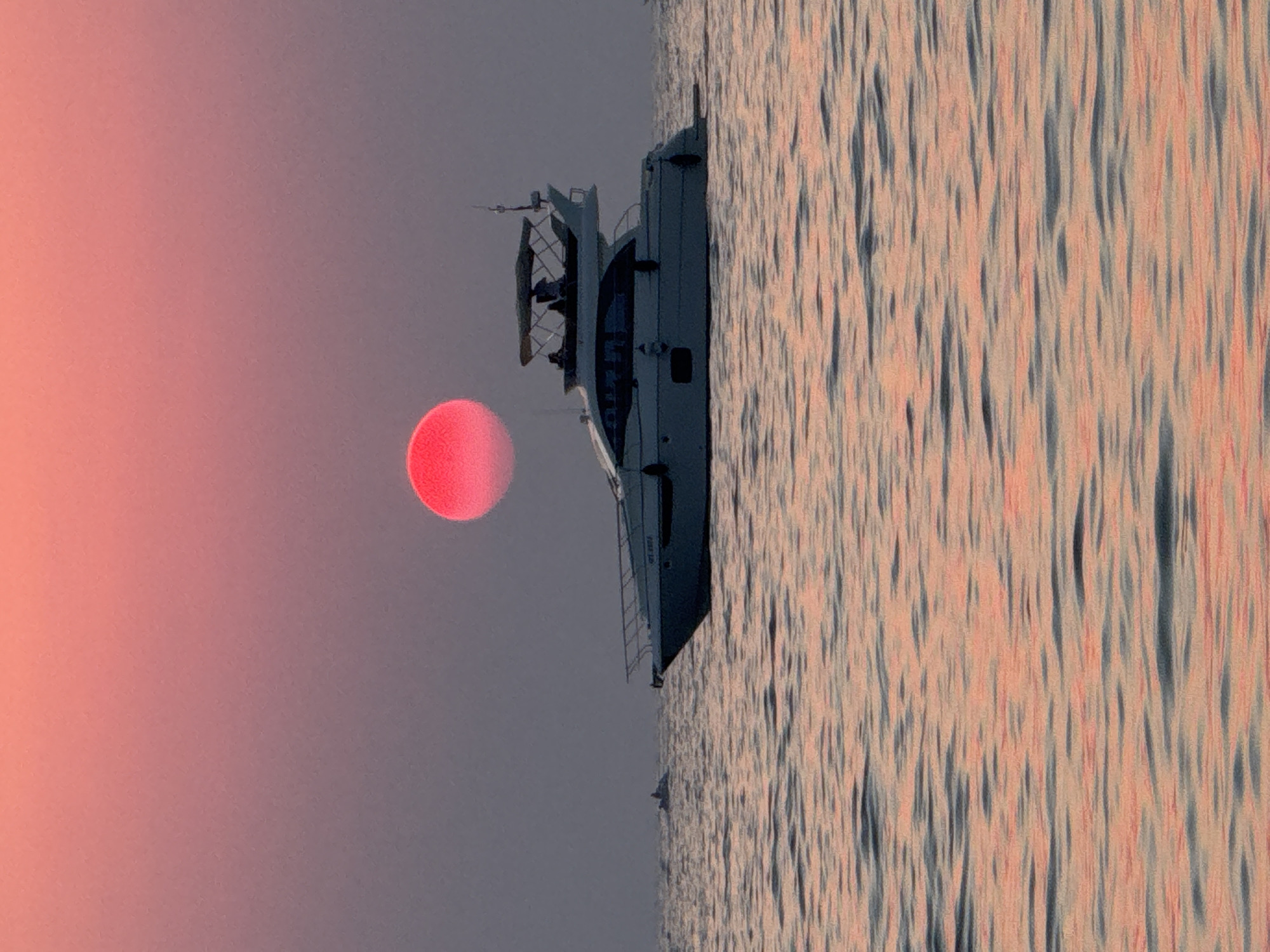 A motor yacht sails on calm waters under a vibrant pink and orange sunset. The sun, a bright red disc, is positioned in the upper center of the frame, slightly to the left. The yacht, white with a dark hull, is moving from left to right across the image. Several people are visible on the deck of the yacht. In the distance to the left, a smaller watercraft, possibly a jet ski, is also visible. The water reflects the colors of the sunset, with ripples creating a textured surface. The overall scene is serene and evokes a sense of leisure and the end of a day.