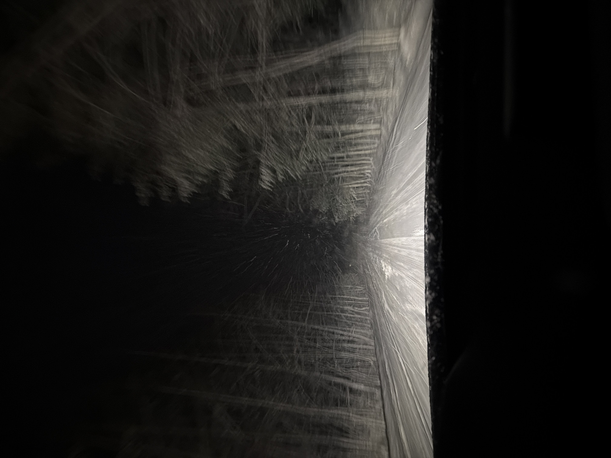 The image shows the interior of a car wash. Rotating brushes are visible through the windshield, covered in water. The scene is dark, with the brushes appearing as blurred white streaks against a dark background. A dark, vertical frame is seen on the right side, likely the car door or window frame.