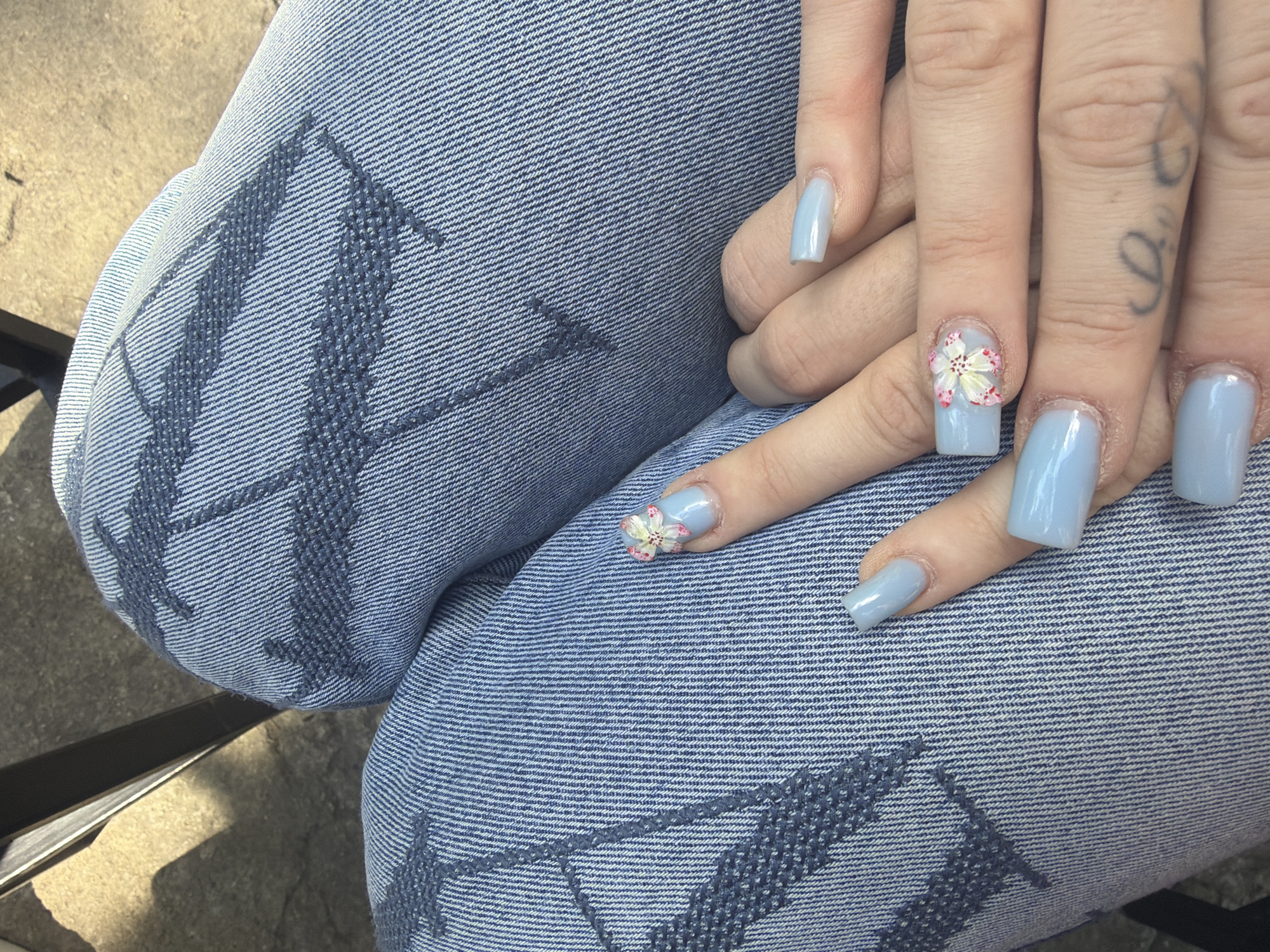 A close-up shot shows a person's crossed legs clad in blue denim jeans adorned with a darker blue stitched pattern resembling large letters. Resting on the jeans are the person's hands with light blue manicured nails. On one of the fingernails is a small, detailed floral design. A tattoo is visible on one of the fingers. The scene appears to be outdoors, with a hint of a dark object and a light-colored surface visible in the periphery. There is no evidence in the image to indicate this photo was taken in Sofia, Bulgaria.