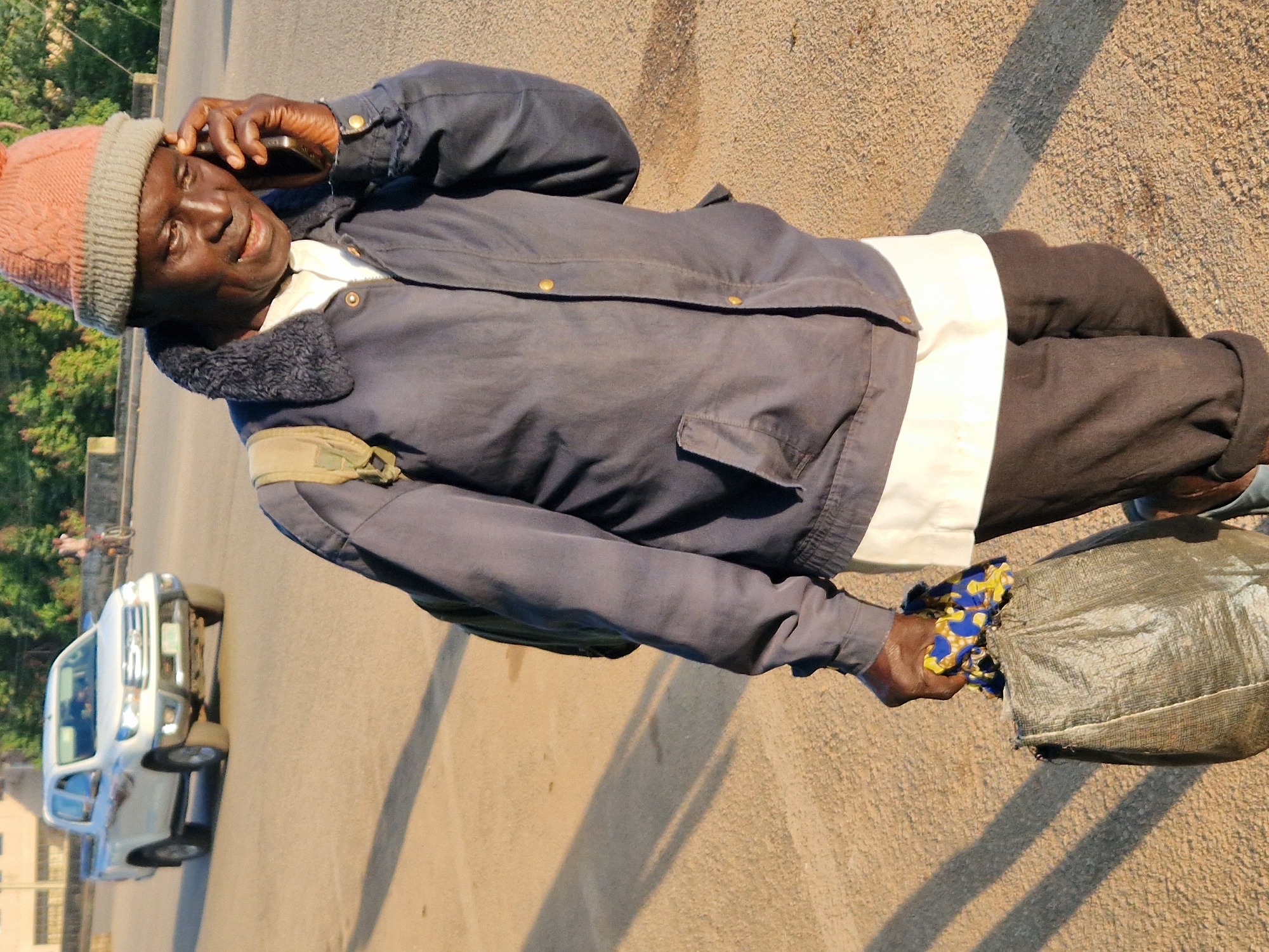 A dark-skinned man in his 60s is walking on a paved road, holding a dark-colored bag in his left hand and talking on a cell phone. He is wearing a dark blue jacket with a fur collar, a white shirt, dark pants, and a knitted hat. He has a light-colored backpack on his shoulders. The man is walking from the left of the frame to the right. In the background, a white pickup truck is driving away from the camera. The sun is low in the sky, casting long shadows on the road. The environment appears to be a street in a city, with some trees and buildings visible in the background.
