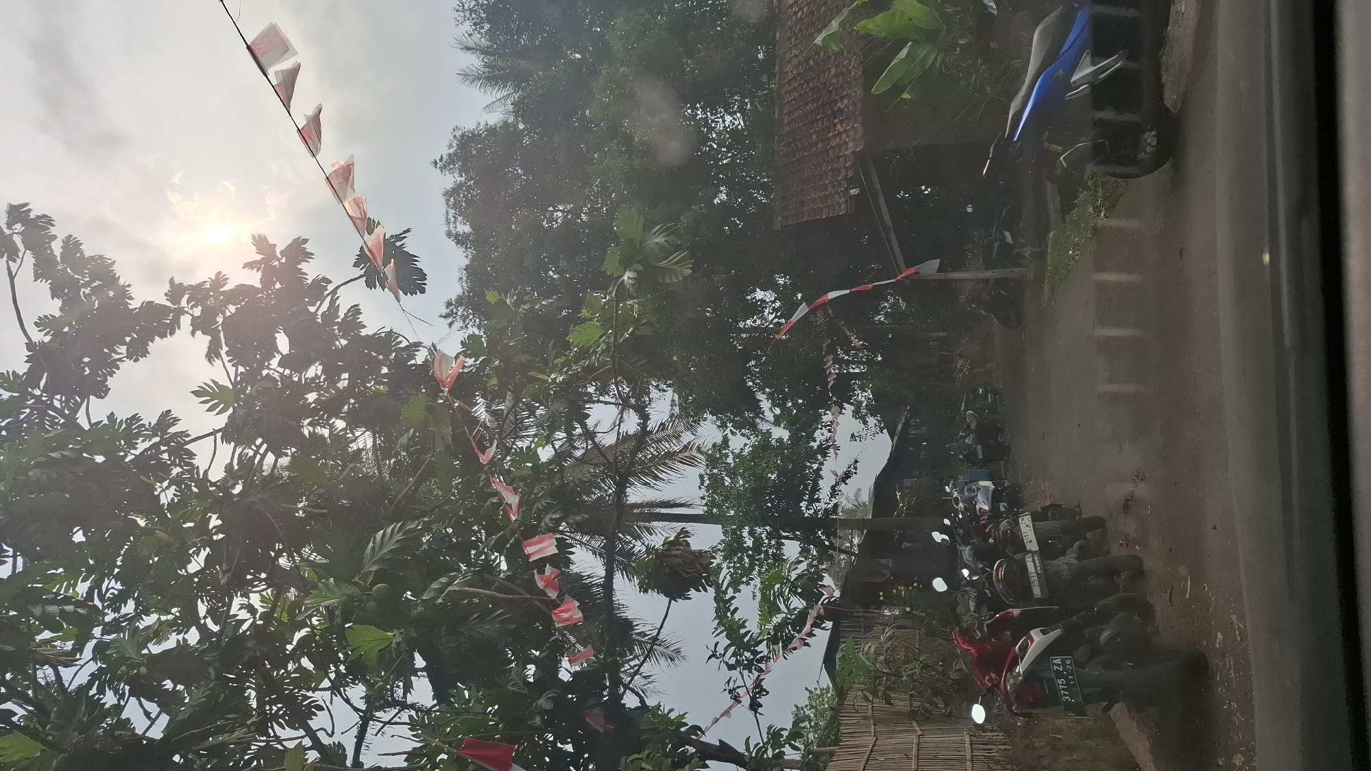 The image depicts a street scene in Sukasari, Indonesia, likely during a local celebration, as evidenced by the red and white Indonesian flags strung across trees and along a pole. The foreground shows the top of a car's dashboard and windshield, through which the scene is viewed. The street is paved and appears wet, suggesting recent rain. A row of parked motorcycles lines the left side of the street, with their headlights illuminated. On the right side of the street, there is a house with a tiled roof and lush green vegetation. The background is filled with dense trees and foliage, with the sun visible through the clouds, casting a bright glare. The overall atmosphere is that of a residential area, possibly a village or a less developed urban setting, with a festive touch.