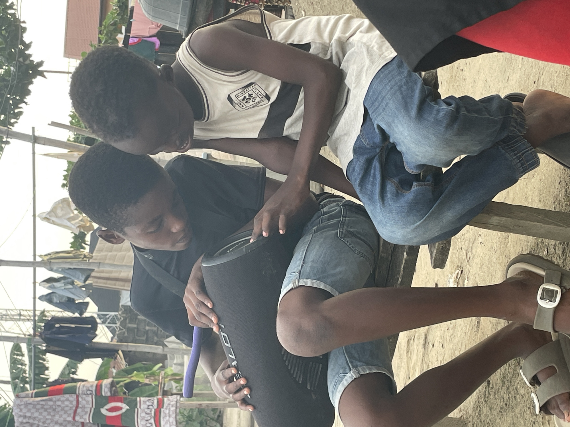 The image depicts two young males outdoors in what appears to be a residential or semi-urban setting in Ado, Nigeria. They are positioned close to each other, seemingly engaged with a portable speaker.
One boy, on the right, is wearing a white sleeveless vest with dark stripes and a logo that reads "MANHATTAN *YORK" within a diamond shape. He is also wearing light blue jeans with elasticized ankles and light grey sandals. He has short, dark curly hair and a slight smile as he looks towards the speaker. He appears to be sitting on a wooden bench or similar structure.
The second boy, on the left, wears a black t-shirt and light blue denim shorts with rolled cuffs. He is holding a black cylindrical portable speaker with both hands. Visible text on the speaker includes "TRONS" (likely part of a brand name like Tronsmart). A purple strap or handle is also attached to the speaker. This boy has short, dark hair and is focused on the speaker.
The scene's background shows various elements suggesting an outdoor, possibly casual living area. There's some vegetation, what looks like wooden posts or fences, and clothes hanging on lines. Notably, one piece of hanging laundry features a distinct pattern with repeated "GUCCI" branding in green, red, and white. The lighting indicates it is daytime. The overall impression is of two boys sharing a moment, possibly listening to music or handling the speaker, in a relaxed outdoor environment.