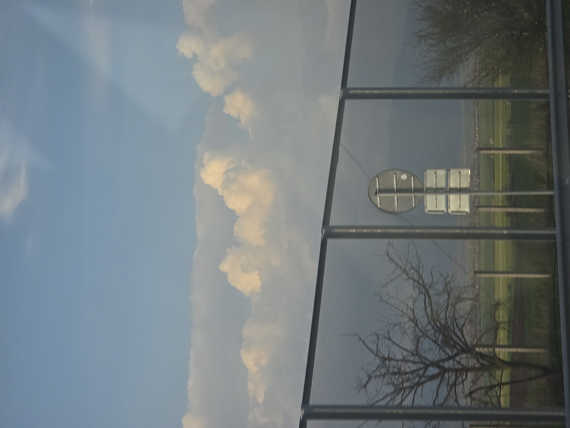 The image shows a view from behind a metal fence. On the left side of the image is a blue sky with white clouds. The right side shows a reflection in the fence, including a road sign, a field, and a tree. The location is not specified in the image, and there is no indication that it is in Chokoba, Bulgaria. There are no people visible in the image.
