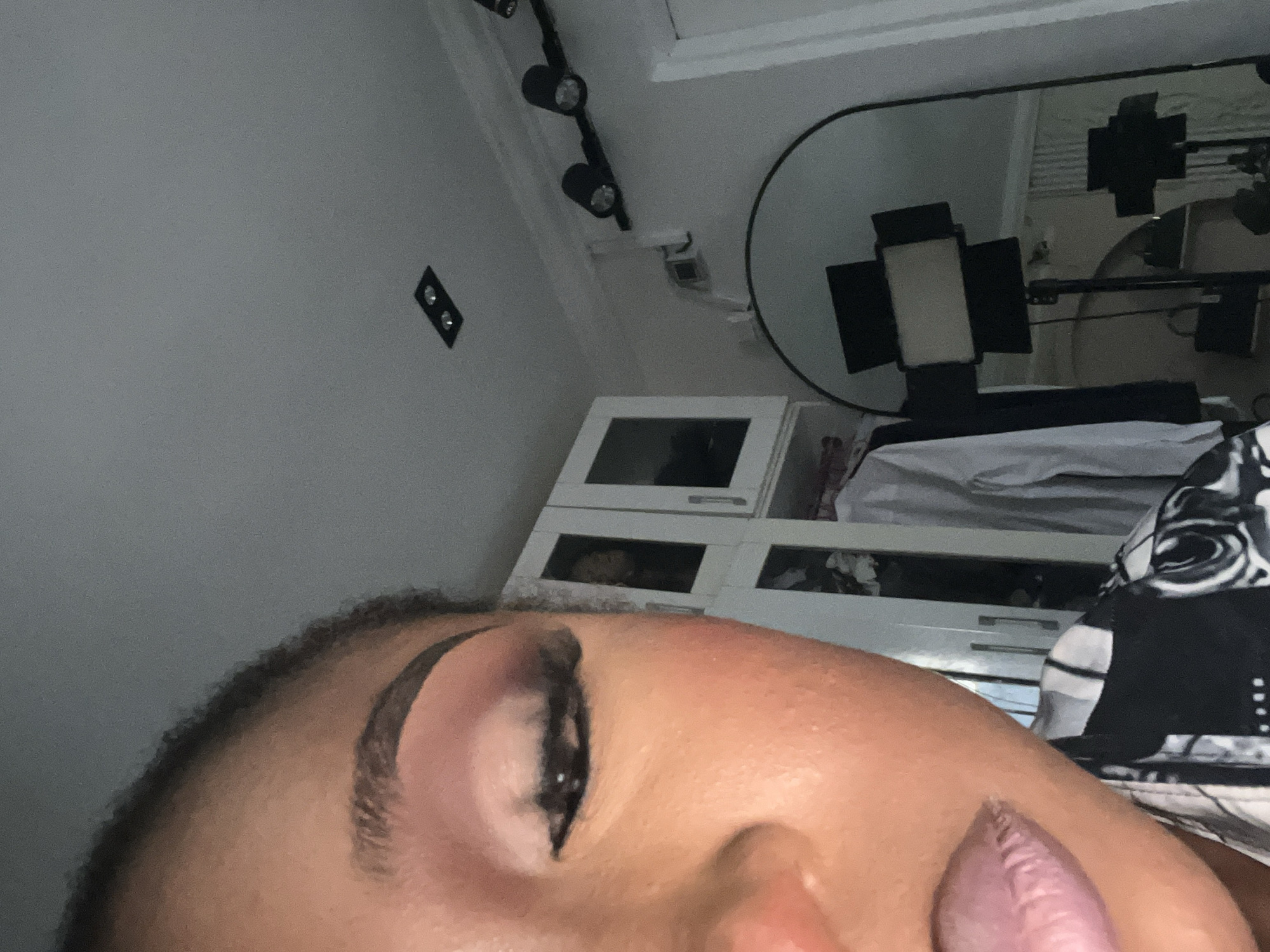 The image shows a close-up, chest-up view of a Black person with their eyes closed, wearing makeup. The person's left eye, eyebrow, and lips are visible. Their skin appears smooth and there are hints of blush on their cheek. They are wearing a black and white patterned shirt.
The background suggests an indoor environment. To the left, there is a white cabinet with glass doors, revealing clothing inside. Hanging on a rack behind it are white shirts. Above the cabinets, there are ceiling lights.
To the right of the person, a large, arched mirror is visible. It reflects studio lighting equipment, including two stands with lights. There are also track lights on the wall above the mirror. The overall impression is of a well-lit space, possibly a room set up for photography or content creation. The scene is clean and organized. There is no indication of the specific location within Abuja, Nigeria, only that it is an indoor residential or studio setting.