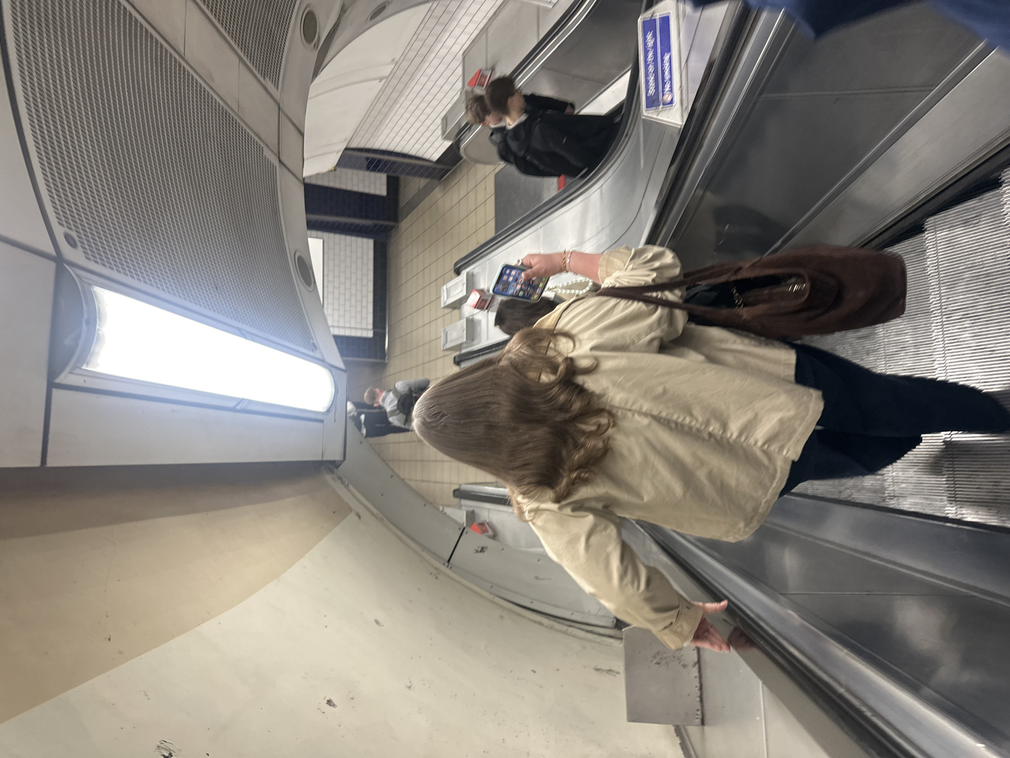 The image captures a scene within a deep underground escalator tunnel, characteristic of a subway or metro system, consistent with the London Underground. The perspective is looking downwards along the escalator, with a slight tilt making the scene appear rotated clockwise.
In the foreground, a person with long, wavy brown hair, wearing a light beige or tan jacket and dark trousers, is descending the escalator. They are viewed from behind, their right hand holding a smartphone, the screen displaying what appear to be app icons. A brown shoulder bag is slung over their left shoulder. Their right hand is positioned near the escalator handrail.
Further down the escalator, another person wearing a dark jacket is visible, also descending. In the background, at the bottom of the escalator, more indistinct figures can be seen.
The escalator features metal ribbed steps and a dark handrail. The curved walls of the tunnel are made of a light-colored material, possibly concrete, with sections of perforated metal ventilation grilles along the upper parts. Long, horizontal fluorescent or LED light fixtures illuminate the tunnel from above and along the walls.
A notable detail is a blue sign with white text, affixed to the side of the escalator structure, next to the handrail. The text on this sign appears to be in Japanese script. The scene depicts typical commuter activity, with individuals utilizing the escalator to traverse the underground station. The artificial lighting suggests an indoor setting, with no indication of the time of day or external weather.