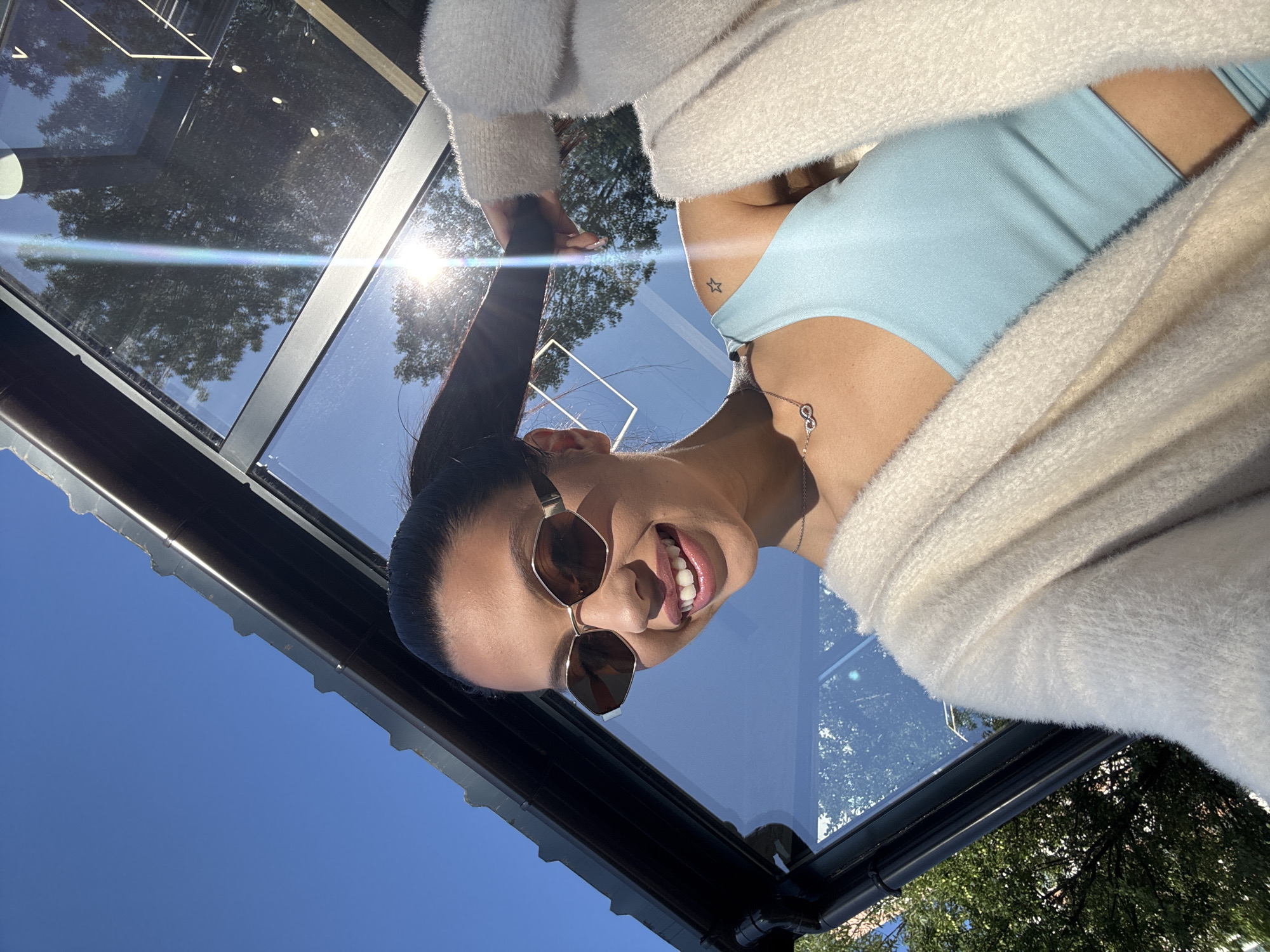A woman is captured in a close-up, outdoor selfie with a bright blue sky and the sun shining. She is wearing sunglasses with geometric frames and a light blue tank top. A fluffy, cream-colored cardigan is draped over her shoulders. Her hair is pulled back in a ponytail, and she is holding a strand of it. She is smiling, and her teeth are visible. A delicate necklace with an infinity symbol is visible on her chest. A small star tattoo is visible on her left shoulder. The background includes reflections of trees and the sky in a glass structure. The scene appears to be in an urban environment, potentially a park or a café with an outdoor seating area. No specific landmarks in Sofia, Bulgaria, are identifiable.