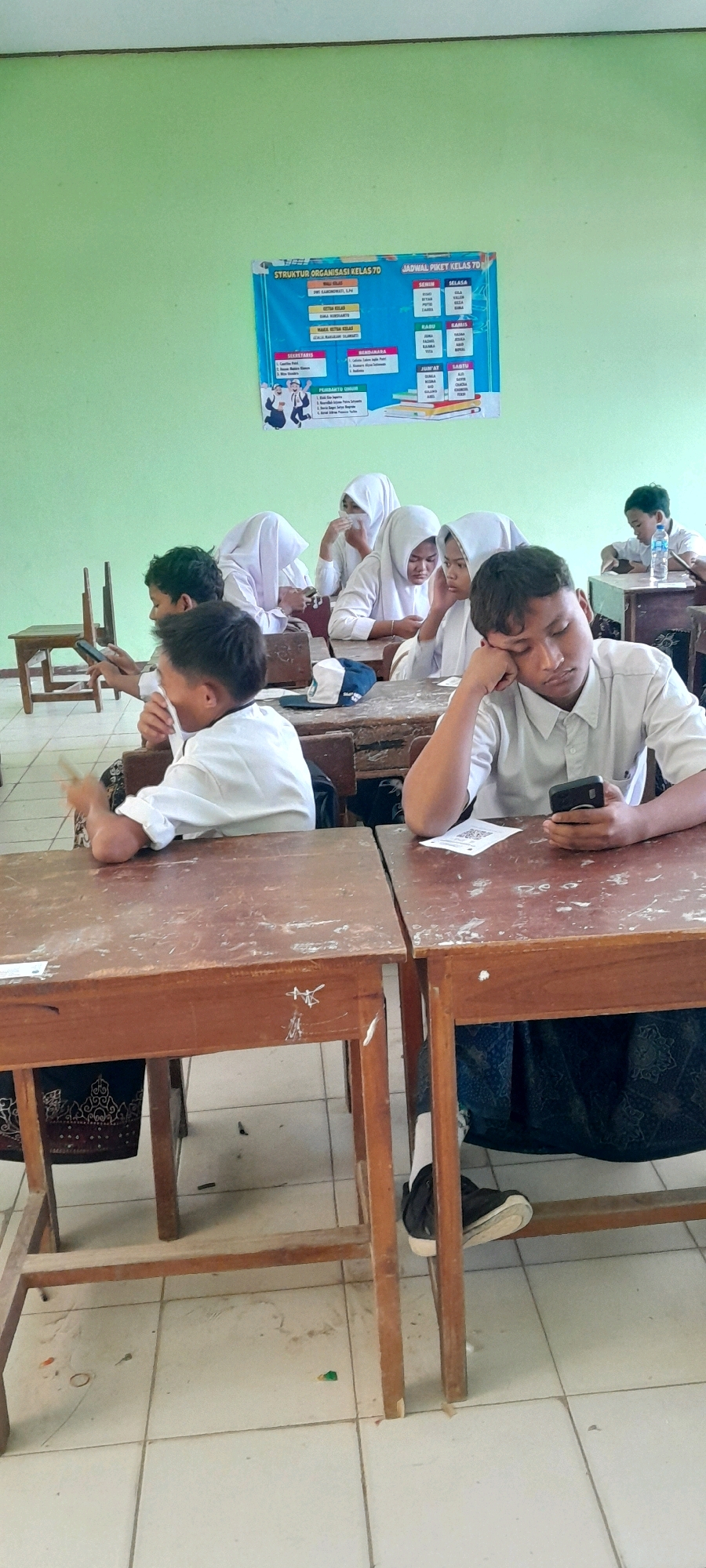 The image depicts a classroom scene in Tahunan, Indonesia, where several students, mostly wearing white uniforms and headscarves, are seated at wooden desks. Some students are engaged with their mobile phones, while others appear to be resting or looking away. A poster is visible on the green wall, containing organizational charts and a schedule. The classroom environment is simple, with tiled flooring and wooden furniture, suggesting a typical educational setting. The lighting appears to be natural, coming from an unseen source.