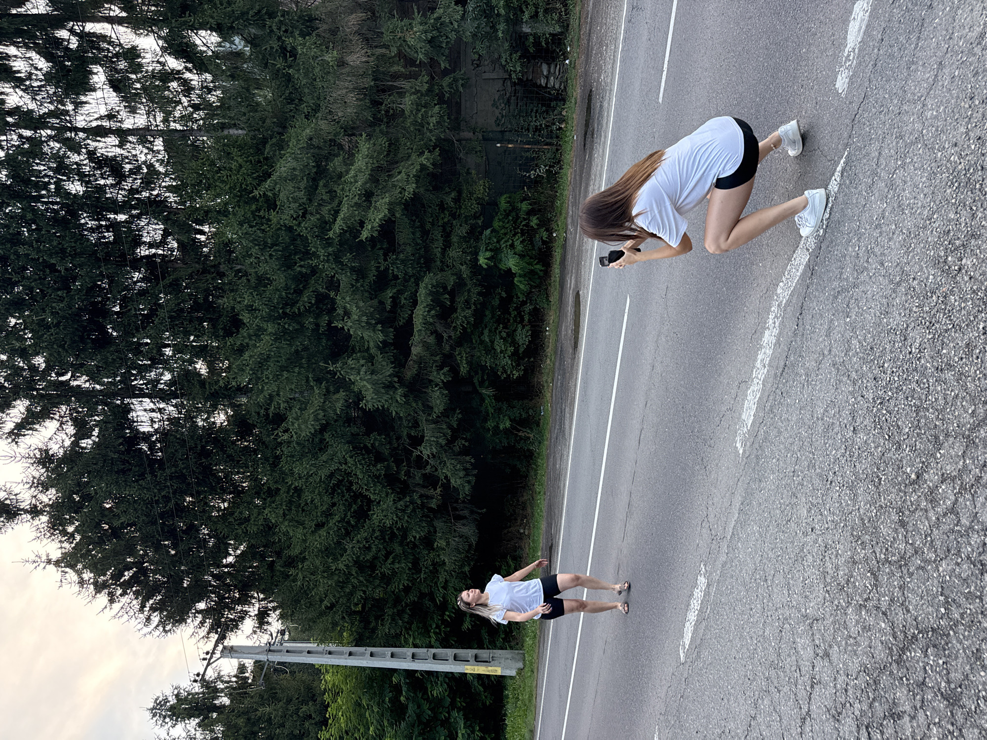 Two women are on an asphalt road in Slănic-Moldova, Romania. The woman on the left is walking towards the right, wearing a white t-shirt and black shorts. The woman on the right is squatting and taking a photo of the woman on the left with a smartphone. She is wearing a white t-shirt, black shorts, and white sneakers. The background is a dense forest of dark green pine trees, with a clear sky above. There is a grey concrete utility pole with wires on the left side of the image.