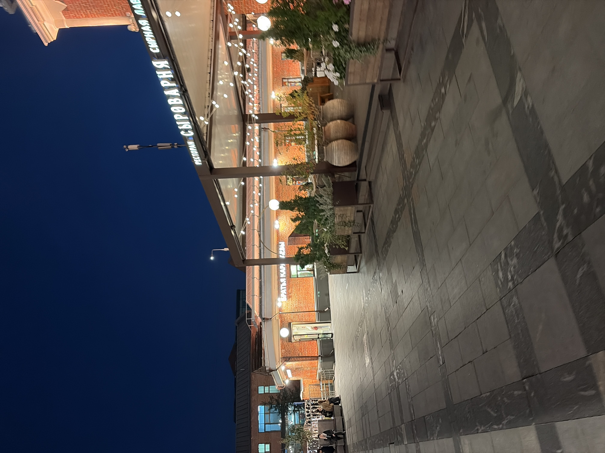 The image captures an outdoor seating area of a restaurant at dusk. The restaurant's facade is made of red brick, and a neon sign in Russian, reading "РЕСТОРАН СЫРОВАРНЯ" (Restaurant Syrovarnya), is visible. The seating area is covered by a canopy adorned with string lights. Large potted plants and decorative vases are placed around the seating area. In the background, a building with large windows and lights is visible. The ground is paved with square tiles. A few people are seated on a bench in the distance, and others are walking. The sky is dark blue, indicating nighttime.