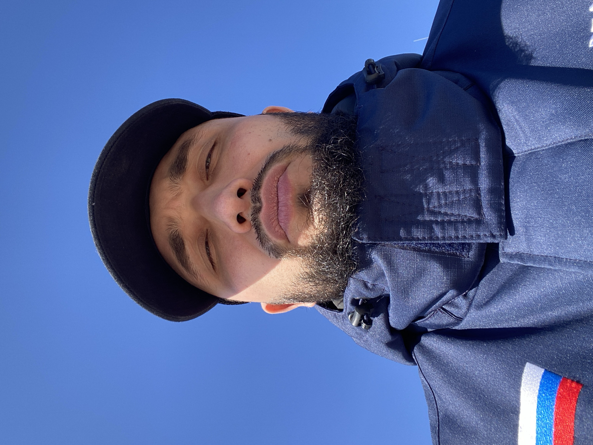 A low-angle, close-up portrait features a man against a clear, bright blue sky in Luzhitsy, Russia. The man, with dark hair, a beard, and a mustache, is looking slightly upwards, with his eyes partially closed or squinting due to the intense sunlight. He is wearing a dark blue baseball cap, the brim of which casts a shadow over the upper part of his face. He is also dressed in a dark blue jacket, which features a prominent patch on the right sleeve. This patch displays three vertical stripes in white, blue, and red, indicative of the Russian flag. The jacket's fabric appears textured and durable. The bright, clear blue sky and strong shadows on the man's face suggest a sunny daytime setting, likely midday. No specific activity is depicted, giving the impression of a moment of pause or a casual outdoor portrait. There is no visible text in the image, apart from the symbolic colors of the Russian flag on the patch.
