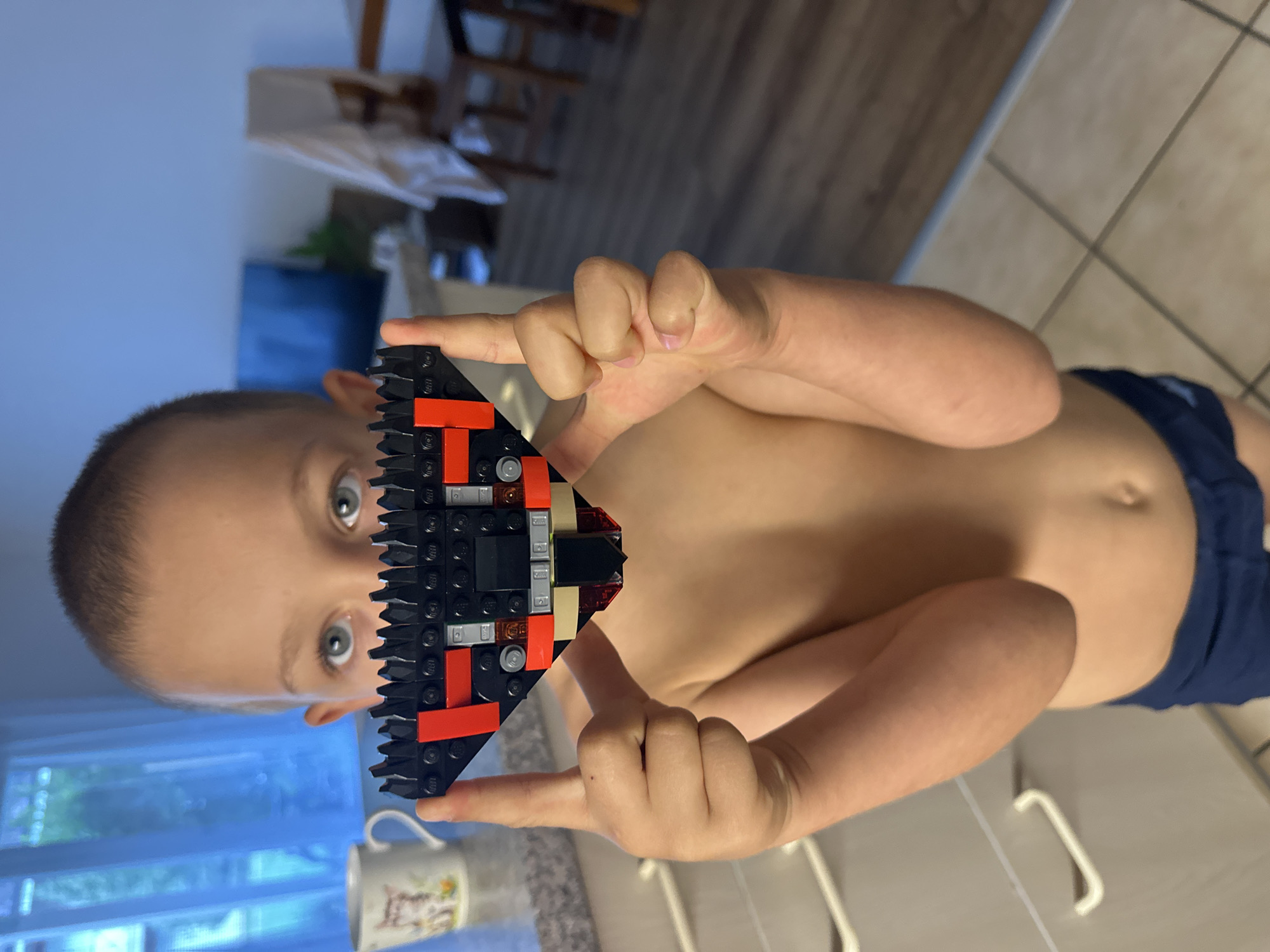 A young boy is holding a black and red LEGO creation up to his face. The LEGO structure is triangular in shape with some silver and tan colored pieces incorporated. The boy's eyes are wide open and looking directly at the camera. He appears to be in a home environment, with kitchen cabinetry visible in the background. The lighting suggests it could be daytime. The image is oriented vertically.