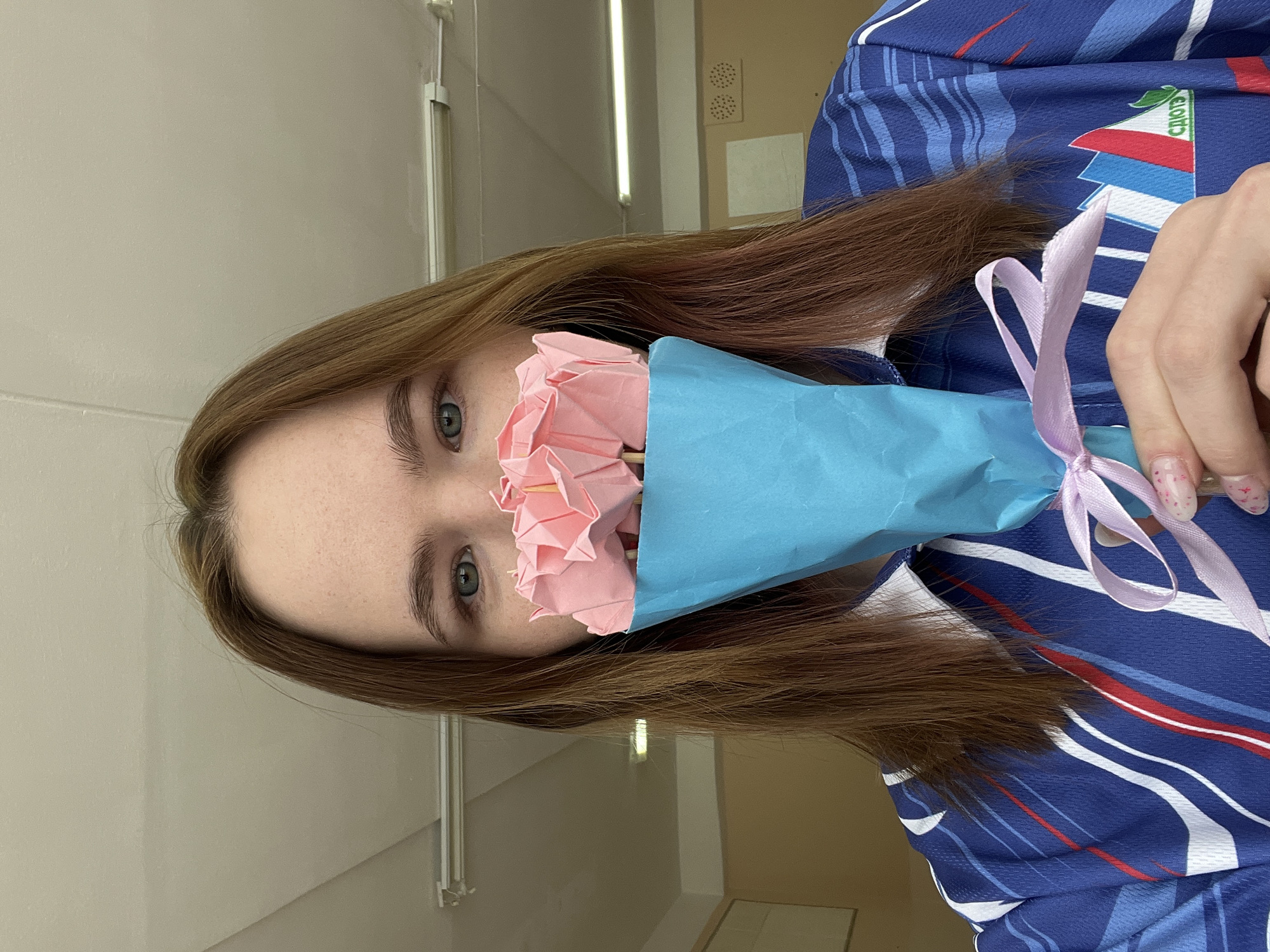 A young woman with light brown hair holds a bouquet of pink paper roses wrapped in blue paper tied with a lavender ribbon. She is wearing a blue jersey with white, red, and navy stripes and a logo featuring the text "СОЮЗ" and a cityscape. The photo was likely taken indoors. The location appears to be a building in Russia based on the inscription on the jersey. Specificity about the city, Berezniki, is not apparent from the image.