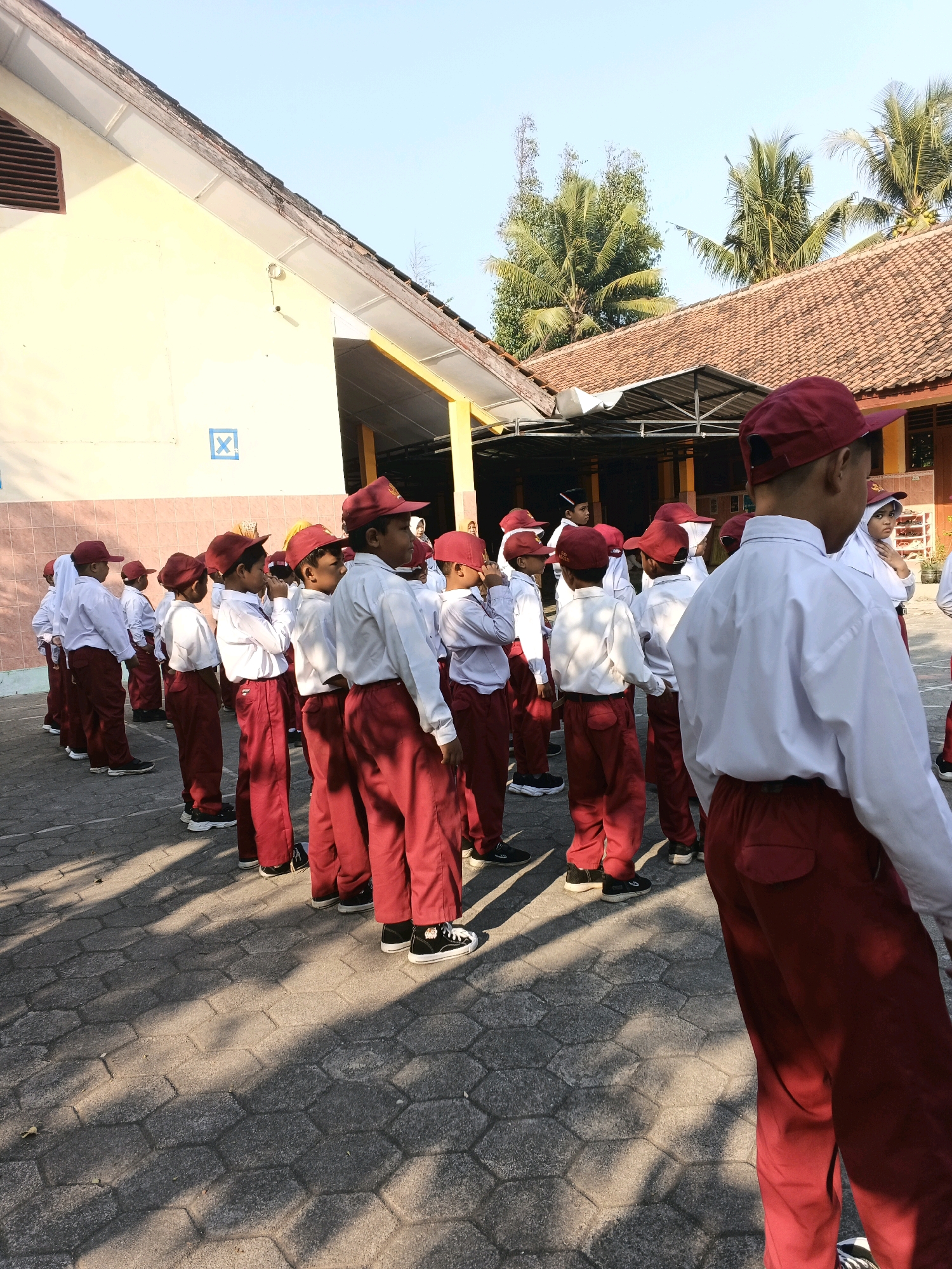 In Glagah, Indonesia, a group of schoolchildren in matching white shirts and red pants, along with red hats, stand in a line outdoors on a paved surface. The children appear to be elementary school age. They are positioned in front of a light yellow building with a red-tiled roof. Sunlight casts shadows on the ground, indicating it is daytime. The building has windows and a covered outdoor area. In the background, palm trees are visible against a clear blue sky. The scene suggests a school assembly or a similar outdoor activity.