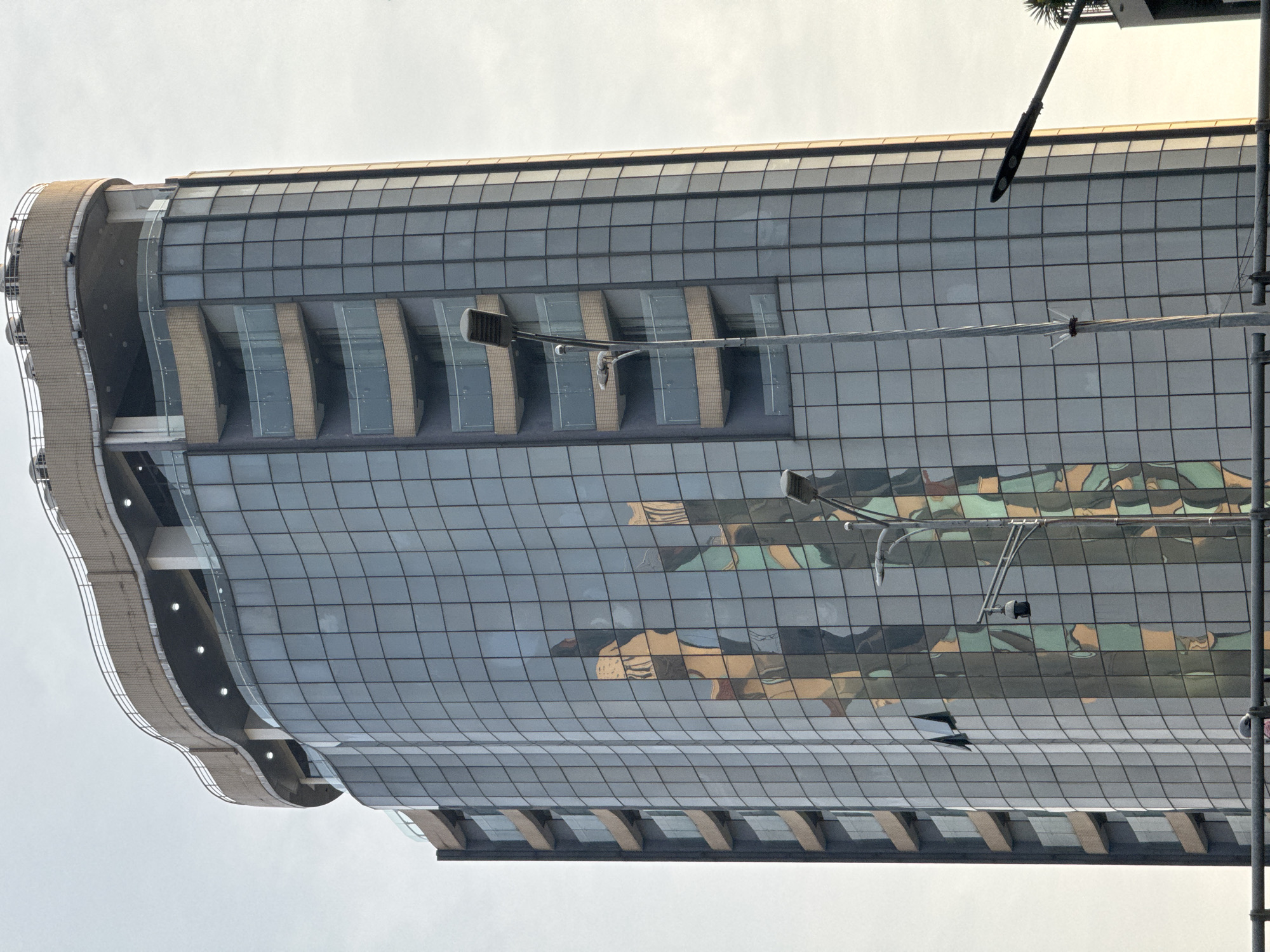 The image shows the exterior of a modern glass-facade building in Hanoi, Vietnam. The building features a curved architectural design with a textured beige facade along its edges and several floors with recessed balconies. The glass panels of the building reflect the sky and surrounding structures, creating a mosaic of colors and shapes. There are streetlights visible, including one with multiple bulbs and a security camera attached to a pole. The sky appears overcast. The orientation of the image is such that the building appears to be tilted, suggesting it was taken from a low angle looking upwards, or the camera was held sideways. The scene is devoid of people and focuses solely on the architecture and its reflections.