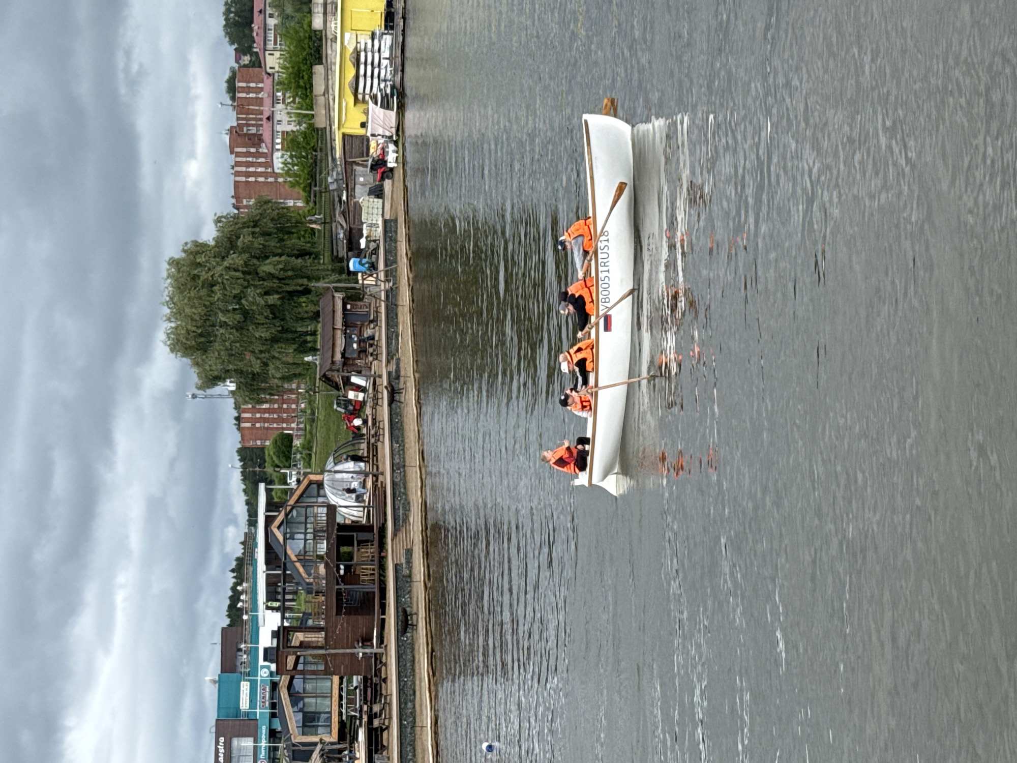 This image captures a daytime scene on the Votkinsk Pond in Votkinsk, Udmurt Republic, Russia, under an overcast sky. In the immediate foreground, a white rowboat bearing the registration "VB0051RUS18" and a small Russian flag decal is occupied by five individuals. All five are wearing orange life vests and are actively rowing with wooden oars on the calm, grey water.
The midground displays a developed waterfront featuring a series of modern wooden structures, including cabins with large windows and angular roofs, alongside a translucent geodesic dome. Wooden jetties and pathways line the shore, where picnic tables are set out. Signs for "Maestra" and "Пятёрочка" (Pyaterochka) are visible on the facades of larger buildings behind these structures. Additional objects on the shore include a small wooden shack, a blue portable toilet, stacks of white bagged materials, a red all-terrain vehicle (ATV), and a yellow building beside stacks of white objects, possibly boat cushions. A large, mature tree with drooping branches is prominent on the right side of the shore.
The background consists of a residential area characterized by multiple brown brick apartment buildings, intermingled with green trees and foliage. A communication tower or antenna structure is visible on the right horizon. Scattered figures are present on the shore near the buildings.