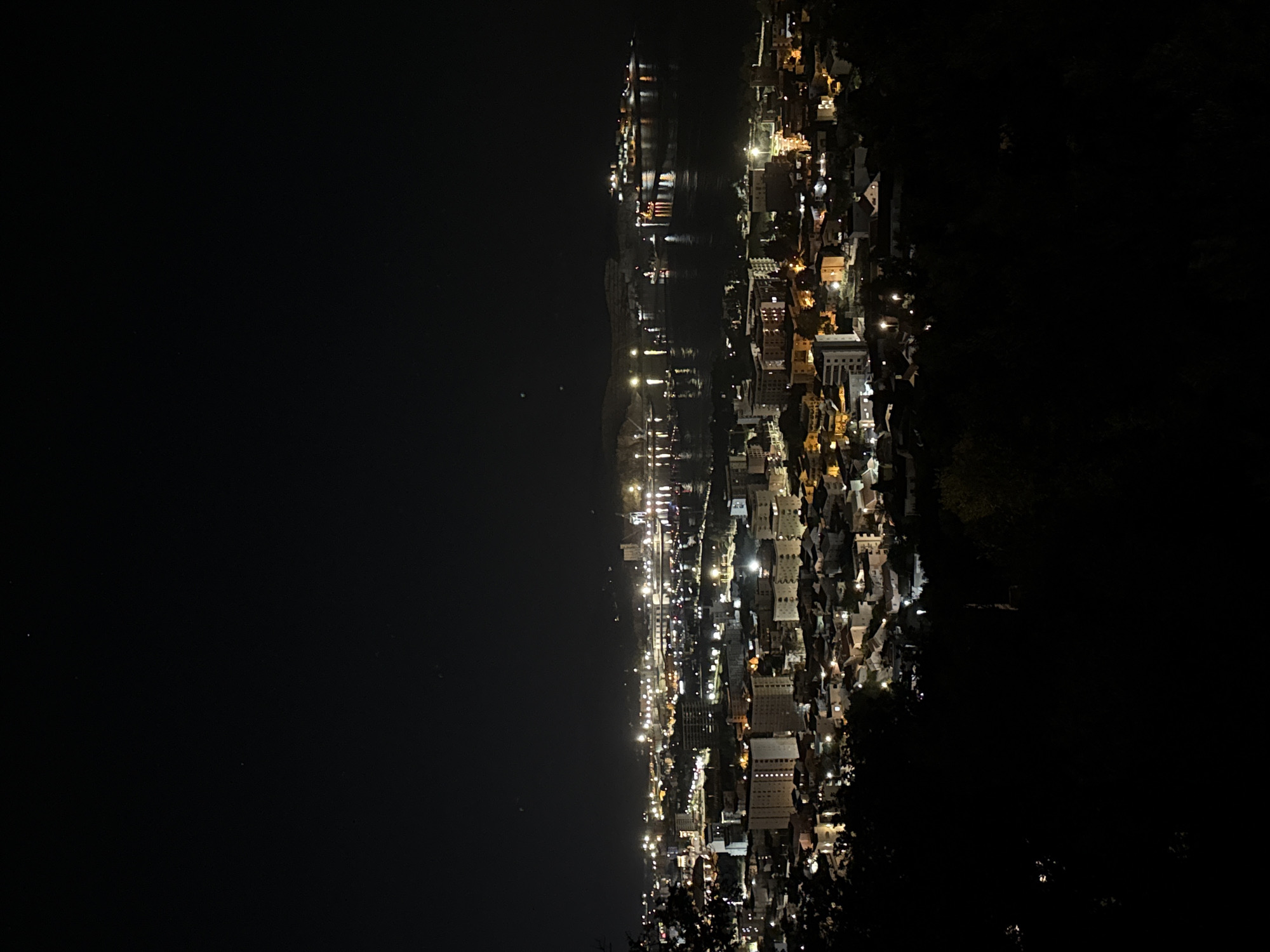 The image depicts an aerial night view of a city, identified as Šušanj, Montenegro. The scene is dominated by the darkness of the night, contrasted by the illuminated urban landscape. The city stretches along the coast, with rows of buildings, roads, and other infrastructure visible due to the streetlights and building lights. A body of water borders the city, reflecting some of the lights. The right side of the frame is enveloped by darkness, suggesting the presence of a hillside or a less developed area.
