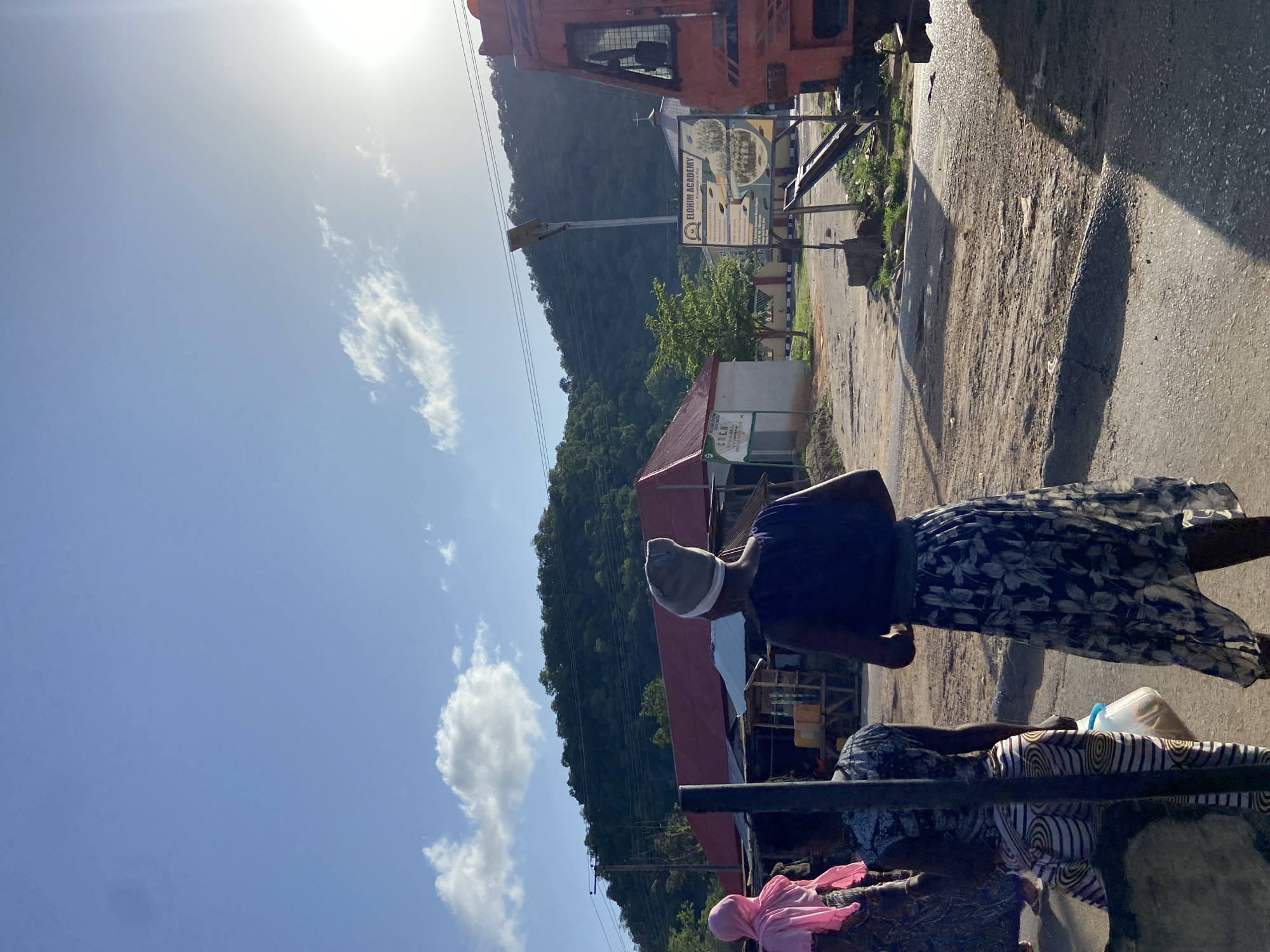 An outdoor shot in Mararaba, Nigeria, shows two Black women on a street near buildings. One woman stands in the foreground wearing a navy shirt, a patterned skirt, and a light-colored hat. Behind her, a second woman sits on what looks like a cart or low trailer, covered in cloth. The second woman is wearing a patterned dress with pink cloth. A clear plastic container sits near her. The background includes a view of buildings with red roofs, a green hill with trees, power lines, and a blue sky with clouds. A partially visible red truck and a school sign are also present. The image is taken during daylight hours with bright sunlight.
