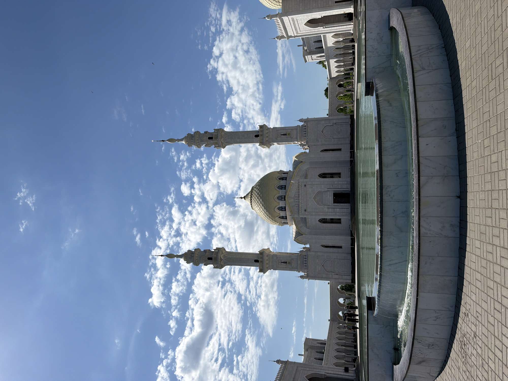 The image shows the White Mosque in Privolzhsky, Russia. The mosque is white with a golden dome and two minarets. A water feature is visible in the foreground. The sky is blue with some clouds. The image is taken during the day. The ground is paved with tiles.