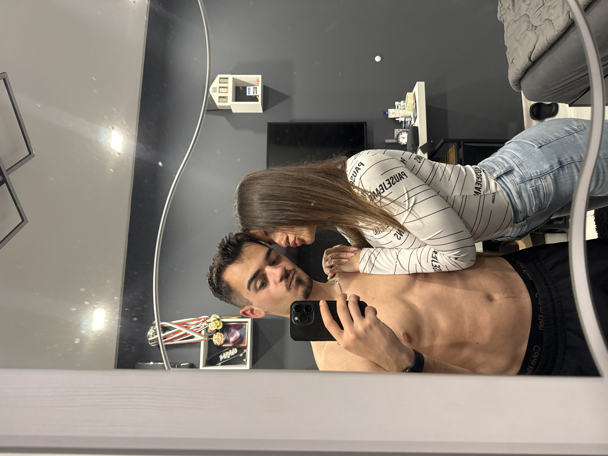 A mirror selfie captures an intimate moment between a man and a woman. The entire image is rotated 90 degrees clockwise from a standard upright orientation, so the subjects appear on their sides.
The man, positioned towards the bottom-center of the frame, is shirtless, revealing an athletic build. He has dark hair, a beard, and a mustache. He holds a black smartphone with a triple camera system in his right hand to take the photo. A dark watch or wristband is visible on his left wrist. The waistband of his black Calvin Klein underwear is visible, displaying the mirrored text "niɘlʞ nivlsƆ".
The woman is situated above and slightly to the right of the man. She has long brown hair and wears a white long-sleeved shirt patterned with black vertical stripes and repeating mirrored text, including "SƎ˥ƎMƎ˥˥M" and "SVAƎd" (likely "EMELLES" and "PAUSE" when unmirrored). She also wears light blue jeans and a ring on her left ring finger. Her head is angled close to the man's, conveying a sense of intimacy and affection.
The reflected background reveals a room with dark grey walls. Visible objects include a black flat-screen television mounted on the wall, a small white multi-compartment shelf unit, and another white shelf holding various smaller items, including what appears to be a clock. A grey upholstered armchair or sofa is partially visible in the top right corner. A small white spherical object is also seen on the dark grey wall. Near the man's reflection, a shelf holds a decorative frame displaying what looks like medals (with red, white, and green ribbons) and a black-framed picture. The lighting is bright and even, suggesting a well-illuminated indoor environment.