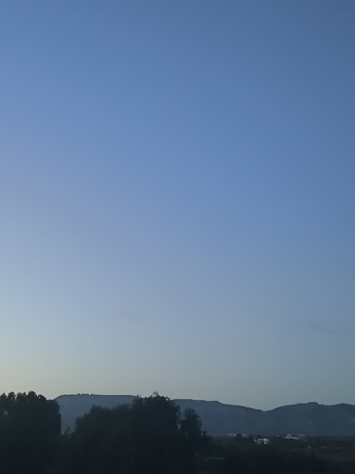 The image displays a panoramic twilight landscape, likely captured from Nicosia, looking north towards the Kyrenia Mountains (Pentadaktylos range). The upper approximately two-thirds of the frame consists of a clear, gradient blue sky, transitioning from a lighter hue at the horizon to a deeper blue overhead. The lower third of the image shows a distant, dark silhouette of a mountain range. Prominently situated on the ridge of the leftmost visible mountain, a series of bright, illuminated white lights are discernible, forming what appears to be a large emblem or structure. In the foreground, below the mountain range and extending across the lower portion of the image, are dark, amorphous silhouettes of trees and vegetation. Faint, scattered artificial lights indicative of urban or suburban development are visible in the mid-ground, particularly towards the right side, nestled within the valley or lower slopes. No individuals, specific actions, or close-up objects are present in the frame.
