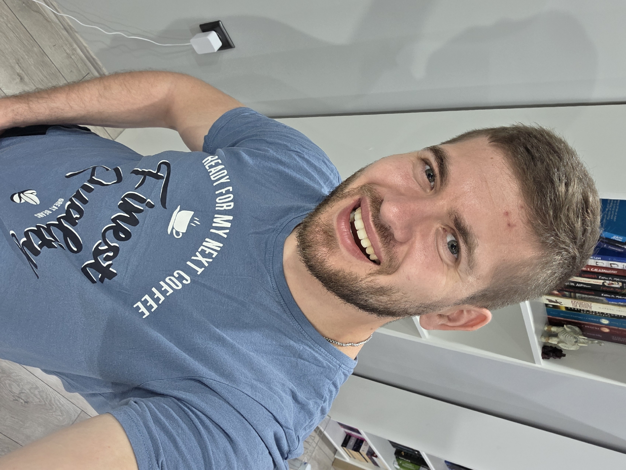 A man with short brown hair and a beard is smiling into the camera. He is wearing a blue t-shirt with the words "Fitness Quality Great Beans READY FOR MY NEXT COFFEE" printed on it. The man is indoors, in front of a white wall and a white bookshelf filled with books and other objects. A power outlet is visible on the wall behind him. The scene is not recognizably located in Yagodovo, Bulgaria.