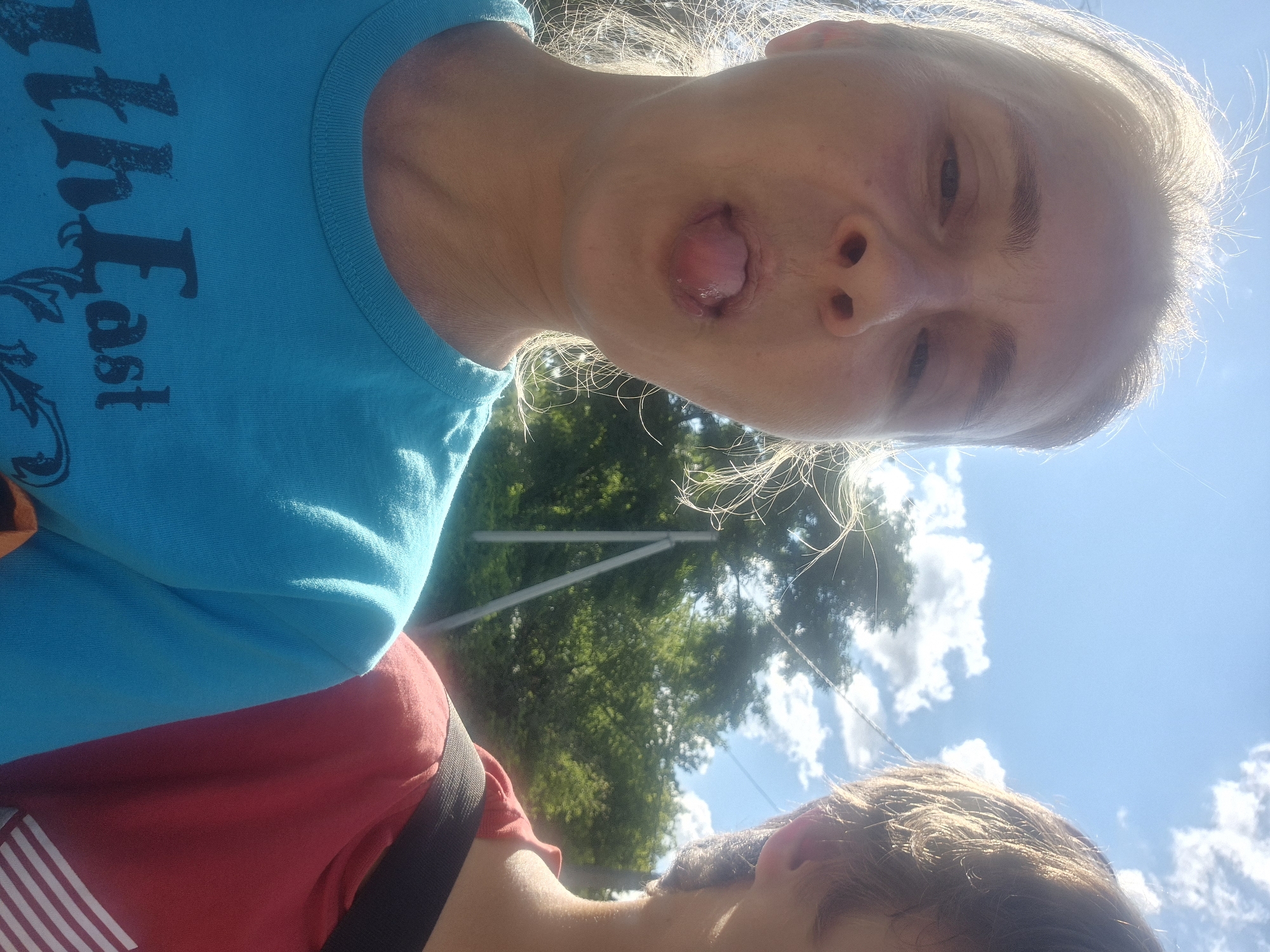 The original image is a close-up, waist-up selfie taken outdoors on a sunny day, likely in Sozonivka, Ukraine, given the information provided. In the foreground, a woman with blonde hair pulled back is sticking her tongue out at the camera. She is wearing a turquoise t-shirt with a dark graphic on the front that appears to read "thEast". To her right, partially in the frame, is a person wearing a red t-shirt with a black strap over their shoulder. The background consists of a bright blue sky with scattered white clouds, green trees, and two light-colored poles in the distance, possibly part of a power line or fence. The lighting suggests it is midday, with the sun shining brightly. The overall scene conveys a casual and playful moment between two individuals.