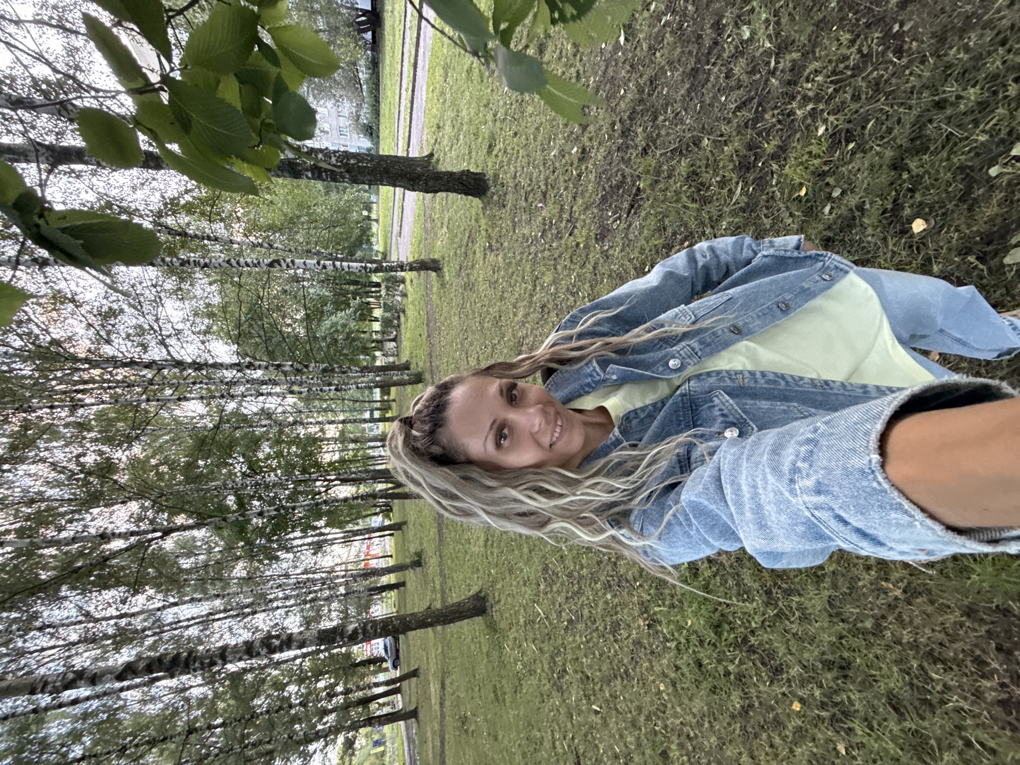 A selfie taken outdoors in a park-like area with birch trees, a paved path, and residential buildings in the background, likely in Rusanovo, Russia. A woman with blonde, curly hair in a high ponytail is facing the camera, smiling. She is wearing a light yellow t-shirt and a blue denim jacket. The ground is covered in grass and fallen leaves. A car is visible in the distance on the paved path.