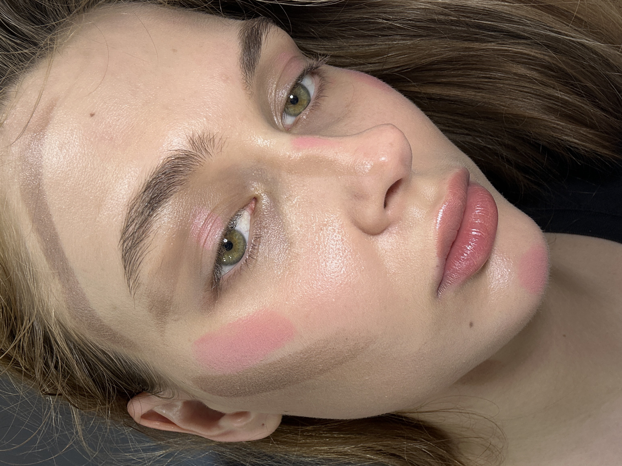A close-up, low-angle shot of a young woman's face. Her skin is light, and she has green eyes. Her eyebrows are light brown and relatively thick. She has noticeable makeup: pink blush, contouring along her cheekbones and forehead, and glossy pink lipstick. Her hair is light brown and appears to be slightly tousled. The background is out of focus and includes some dark fabric. The location, Krasnoyarsk, Russia, cannot be determined from the image.