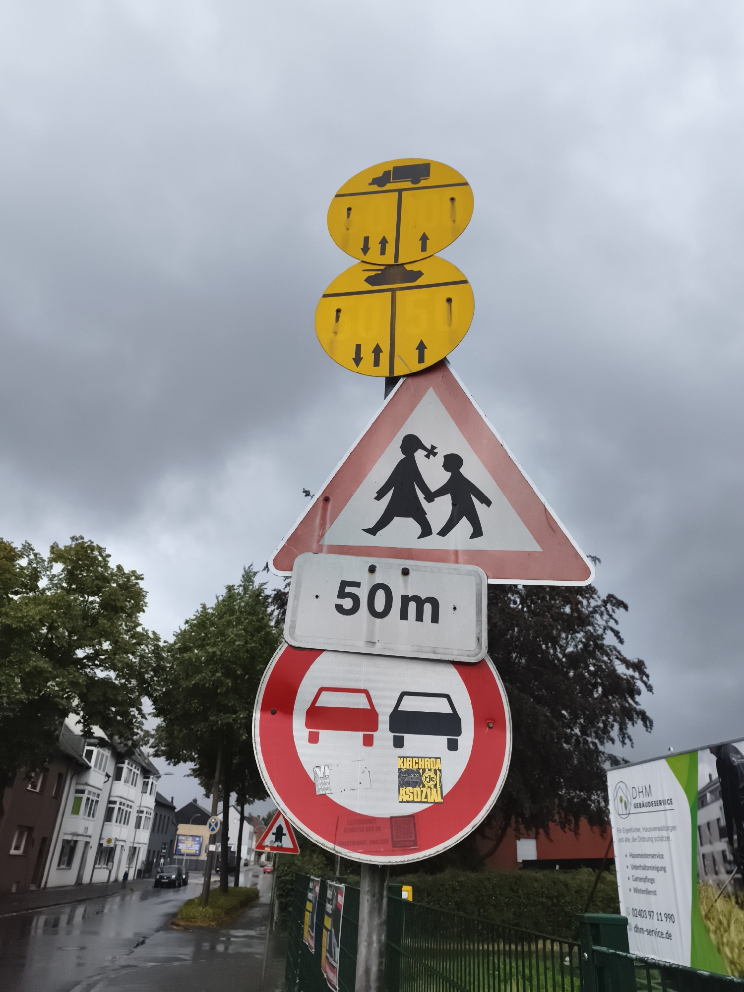 The image shows a street scene in Eschweiler, Germany, on a cloudy day. Multiple traffic signs are prominently displayed on a pole. The topmost signs are two stacked yellow circles with black symbols indicating truck and tank restrictions, with arrows pointing upwards. Below these is a triangular sign with a red border and a white interior, depicting two children walking and holding hands, signaling a school zone or pedestrian crossing. Beneath that is a rectangular sign with "50m" printed on it. The largest circular sign is a red and white prohibition sign with silhouettes of cars, indicating a no-entry zone for vehicles. A sticker with the text "KIRCHROA SOZIAL" is affixed to this sign. To the right, a white banner with green accents advertises "DHM GEBÄUDESERVICE" and lists services such as "Hausmeisterservice," "Unterhaltsreinigung," "Gartenpflege," and "Winterdienst," along with a phone number and website. The street itself is wet, suggesting recent rain. Residential buildings with multi-story facades line the left side of the street, and trees are visible in the background. A black car is driving on the wet street.