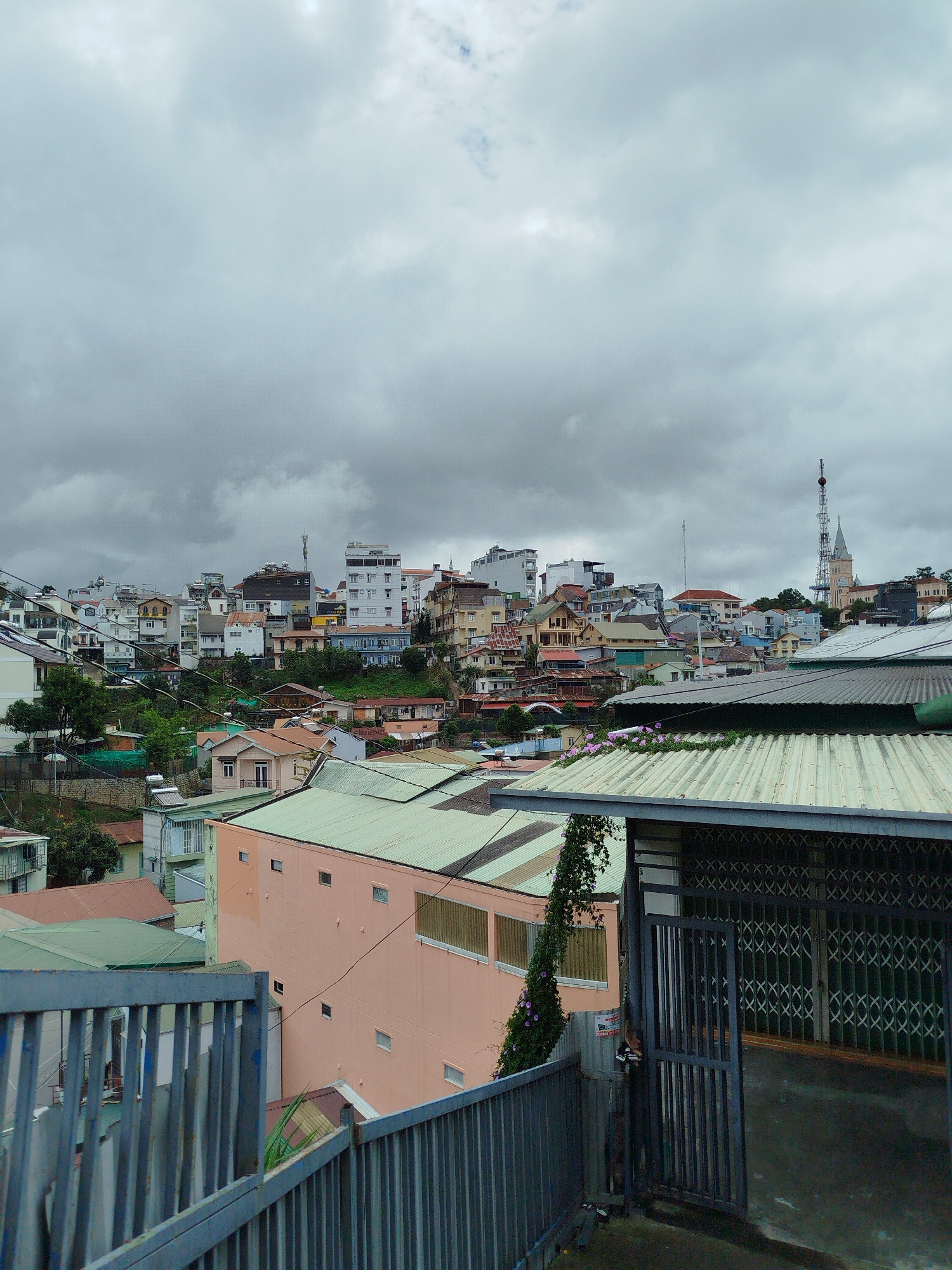 This media file presents a panoramic view of a densely built urban area, likely a city or town situated on hilly terrain, consistent with the landscape of Da Lat, Vietnam. The overall atmosphere is subdued due to an overcast sky filled with heavy grey clouds, suggesting a cloudy day.
In the immediate foreground, a metal railing, possibly part of a staircase or balcony, cuts across the bottom left. To the right, a corrugated metal roof dominates, partially covered by a creeping vine with small purple flowers. A dark grey metal gate, partially open, is visible on the far right, leading into what appears to be a covered entryway. A wall behind the gate shows a faint red banner with a phone number: "0908 316 63".
The midground reveals a sprawling collection of houses and multi-story buildings, stacked closely together on the sloped hillsides. The architecture is varied, with buildings painted in pastel shades like peach, white, and light blue, often featuring contrasting roofs of green corrugated metal or reddish-brown tiles. Green trees and dense foliage are interspersed among the structures, breaking up the urban fabric. Numerous power lines or utility cables crisscross horizontally across the scene.
Further in the background, more buildings extend up the hills, culminating against the grey sky. On the distant horizon to the right, a tall communication tower with an antenna stands prominently, adjacent to a church steeple, adding a distinctive landmark to the cityscape. The time of day appears to be daytime, but the heavy cloud cover gives the scene a diffused, soft light. No specific human activity or people are visible, lending a quiet, contemplative mood to the image of this bustling hillside community.