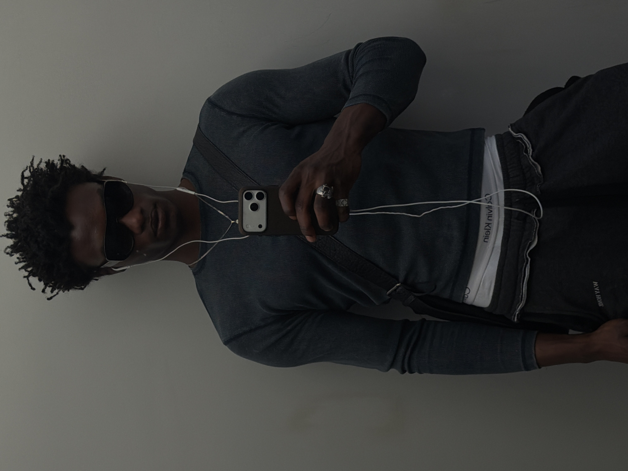 A man with dark skin and curly hair is taking a selfie with his phone. He is wearing sunglasses, a gray long-sleeved shirt, and dark pants with a white Calvin Klein waistband visible. He has headphones in his ears and rings on his fingers. The background is a plain gray wall. The man's body is oriented vertically and at an angle. There is no indication of London or any other location.