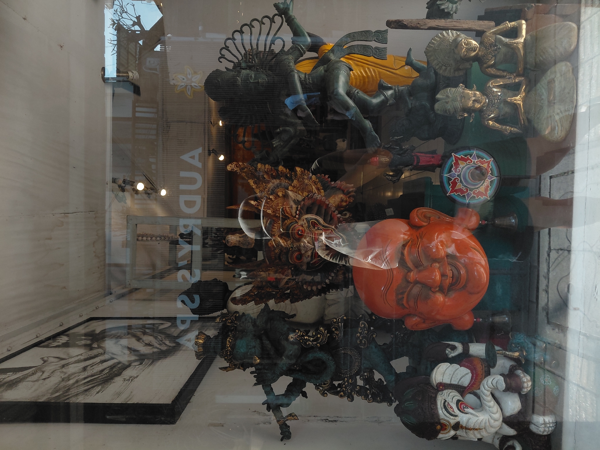 The image shows a reflection in a shop window displaying various statues and decorative items. The shop's sign, reflected in the glass, reads "AUDREY SPA" backwards. On the left side, a statue of Ganesha is visible. To its right, a large, smiling orange mask or head is prominently displayed. Behind these, several other statues are arranged, including a dark-colored figure playing a flute and another in a dynamic pose with a fan-like headdress. Two small, golden statues of people in prayer are positioned in the foreground on the right. The overall impression is of an eclectic collection of cultural artifacts, likely for sale. The backdrop of the reflection shows an outdoor scene with trees and structures.