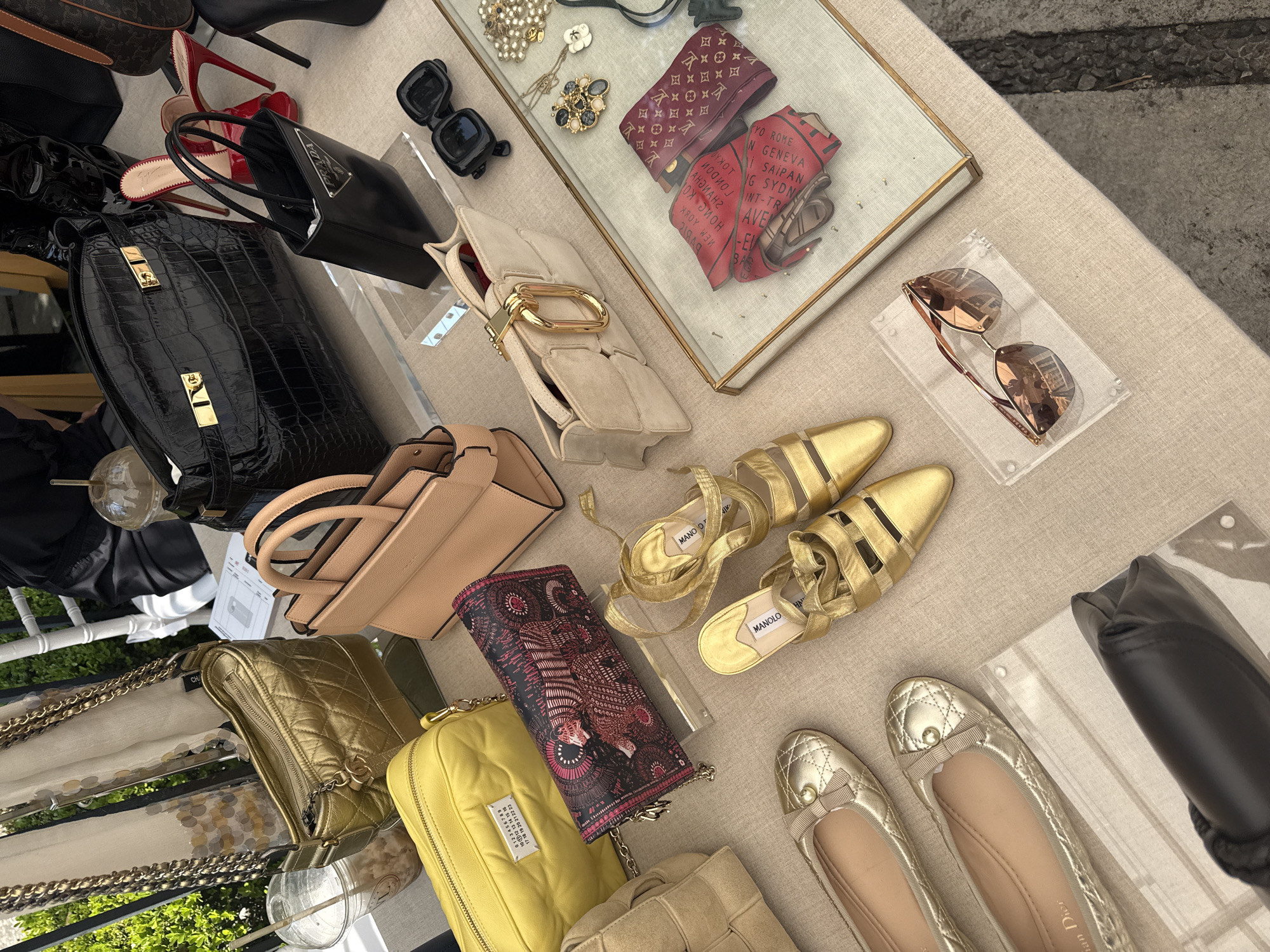The photograph depicts an array of luxury goods laid out on a table, likely for sale or display. The assortment includes handbags of varying styles and colors, ranging from black crocodile-patterned leather to quilted gold and yellow designs, alongside shoes such as gold Manolo Blahnik strappy sandals and silver ballet flats with bows. A beige clutch with a gold clasp is also present. Accessories like sunglasses, including a pair of black square frames and another pair with brown lenses, are scattered among the items. Jewelry, including pearl necklaces and ornate brooches, is arranged in a glass case. The items are arranged on a beige-colored tablecloth, and some are displayed on clear acrylic stands. The setting appears to be outdoors, possibly at a market or private sale, with a glimpse of greenery visible in the lower left corner. The lack of identifying information precludes pinpointing the precise location within Mexico City, Mexico.