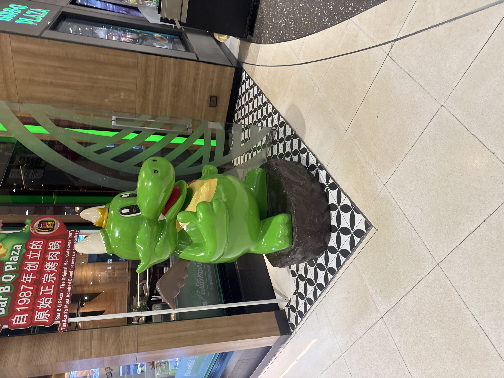 The image shows a green, cartoonish crocodile statue wearing a gold crown and a yellow bib, standing on a dark brown, rock-like pedestal. The statue is positioned in front of a restaurant with a glass door and wooden paneling. A sign is visible to the left of the statue, with text in Chinese and English stating "Bar B Q Plaza - The Original Moo Krata since 1987, Thailand's Most Admired Brand for over six consecutive y". The floor in front of the restaurant has a black and white geometric tile pattern, which transitions to a light-colored tiled floor. The background shows the interior of the restaurant, with tables and chairs visible through the glass. The overall scene appears to be an outdoor area in a commercial setting, likely a shopping mall or plaza in Wichit, Thailand. There are no people visible in the image.