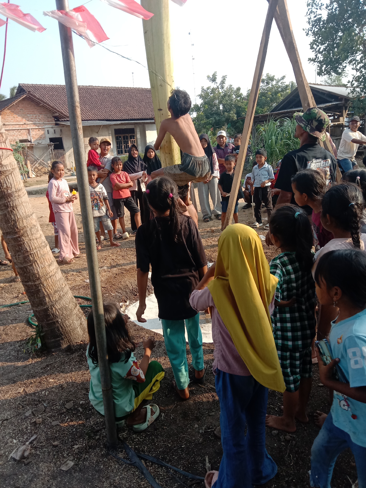 The image depicts an outdoor gathering of people, predominantly children and some adults, in what appears to be a village or rural setting. The scene is characterized by a wooden structure that resembles a game or competition apparatus, with a person suspended from it. Red and white Indonesian flags are visible, suggesting a national celebration or event. Several children are standing and observing the person on the structure, while others are seated or grouped around. Adults are also present, some interacting with the children and others observing the activity. The ground is rough and unpaved, with scattered debris. In the background, there is a building with a tiled roof and vegetation. The overall atmosphere suggests a community event or a traditional game being played in Utama Jaya Mataram, Indonesia.