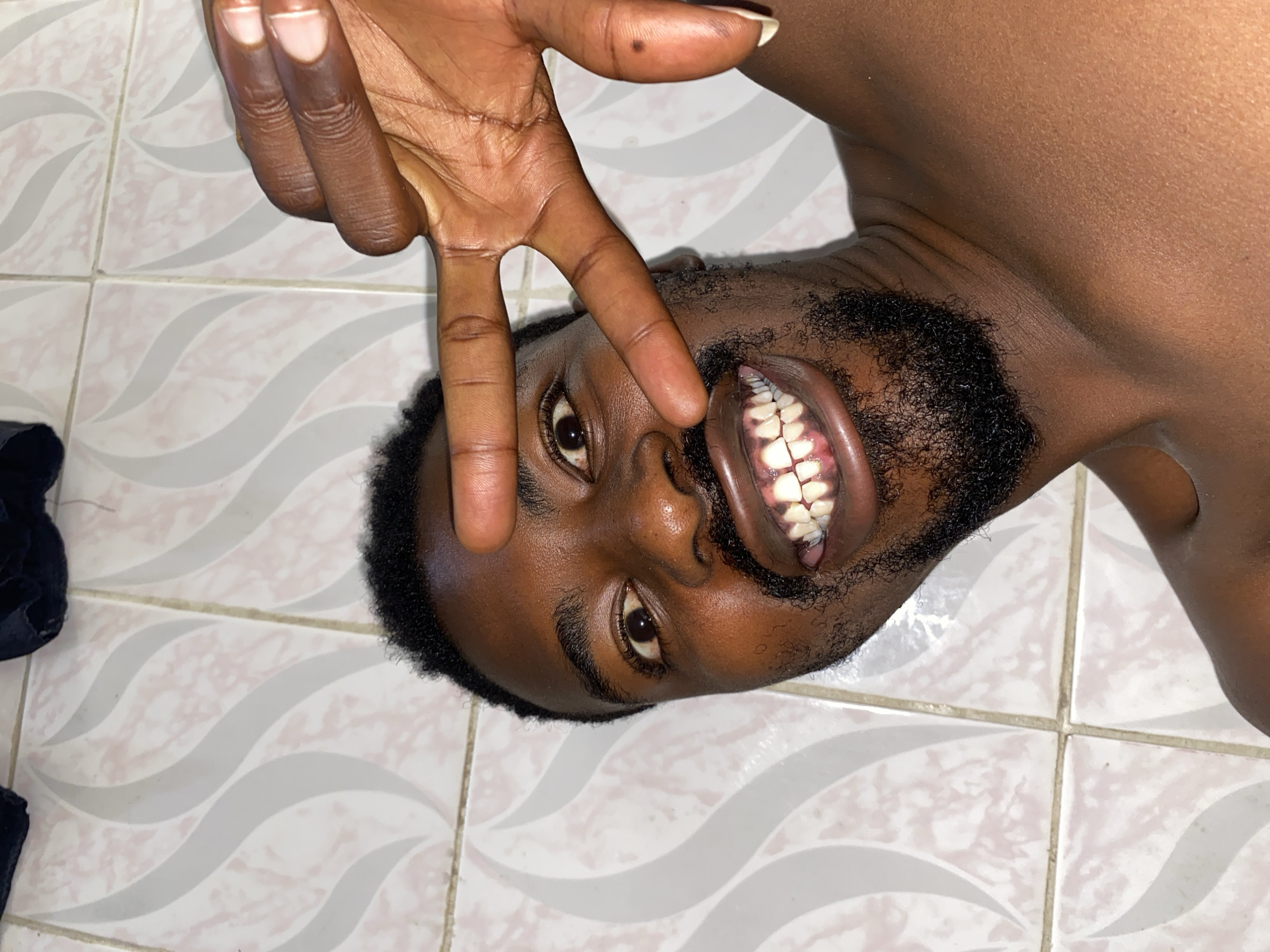 A dark-skinned man lies on his back on a tiled floor, with his head tilted to the side. He is holding his hand up near his face, making a gesture with his fingers. The man has a beard and his mouth is open, revealing his teeth. The tiled floor has a wave pattern in gray and white. Part of a dark object is visible in the lower-left corner. The location is Barakin Akawo, Nigeria.