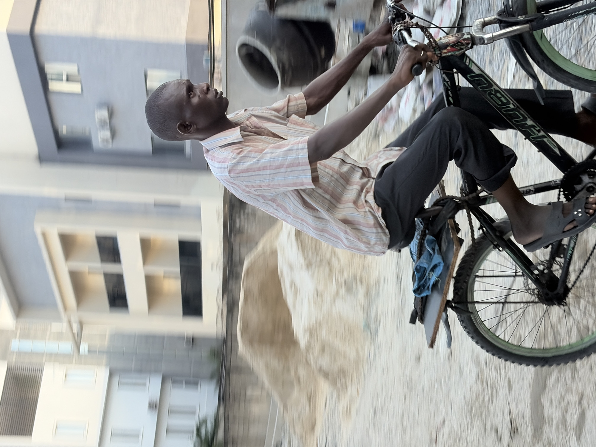 In Moba, Nigeria, an individual rides a black mountain bike on a dusty or sandy road, wearing a light-colored, striped shirt and dark pants. The rider wears grey sandals. A blue cloth is attached to the bike frame. Behind the rider, there is what appears to be construction material, and multi-story buildings are visible in the background.