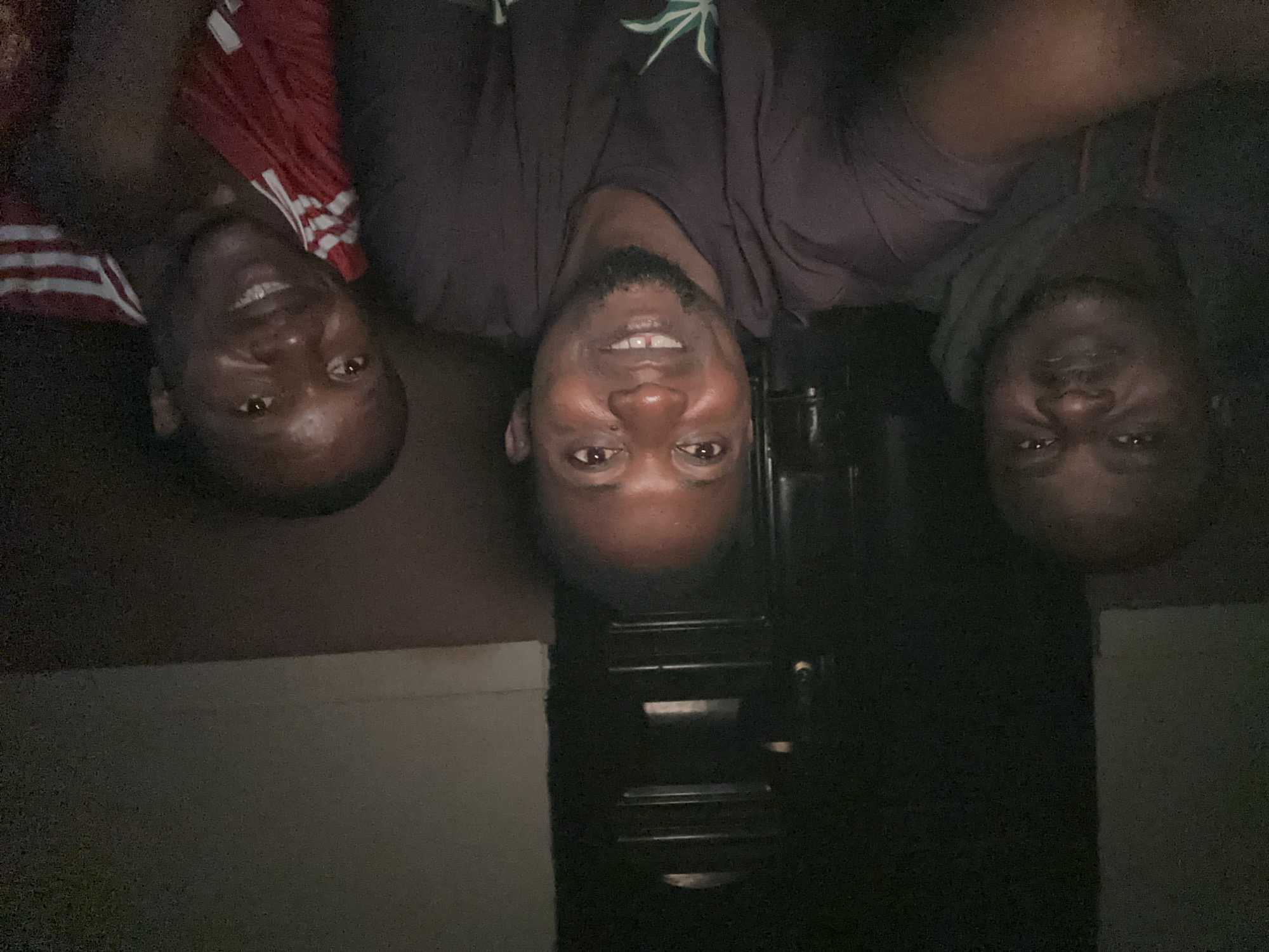 Three men are captured in a selfie. The man in the center, wearing a dark t-shirt with a faint green graphic, is smiling with his mouth slightly open, revealing teeth. He has a goatee. To his left, a man wearing a dark hoodie and a grey t-shirt is visible. To the right, a man in a red shirt with white stripes on the sleeves is smiling. The background is dark and appears to be indoors, with a textured wall on the right and a dark vertical structure with horizontal lines behind the central figure. The lighting suggests a dim environment, possibly at night. The image appears to have been taken in Narayi, Nigeria.