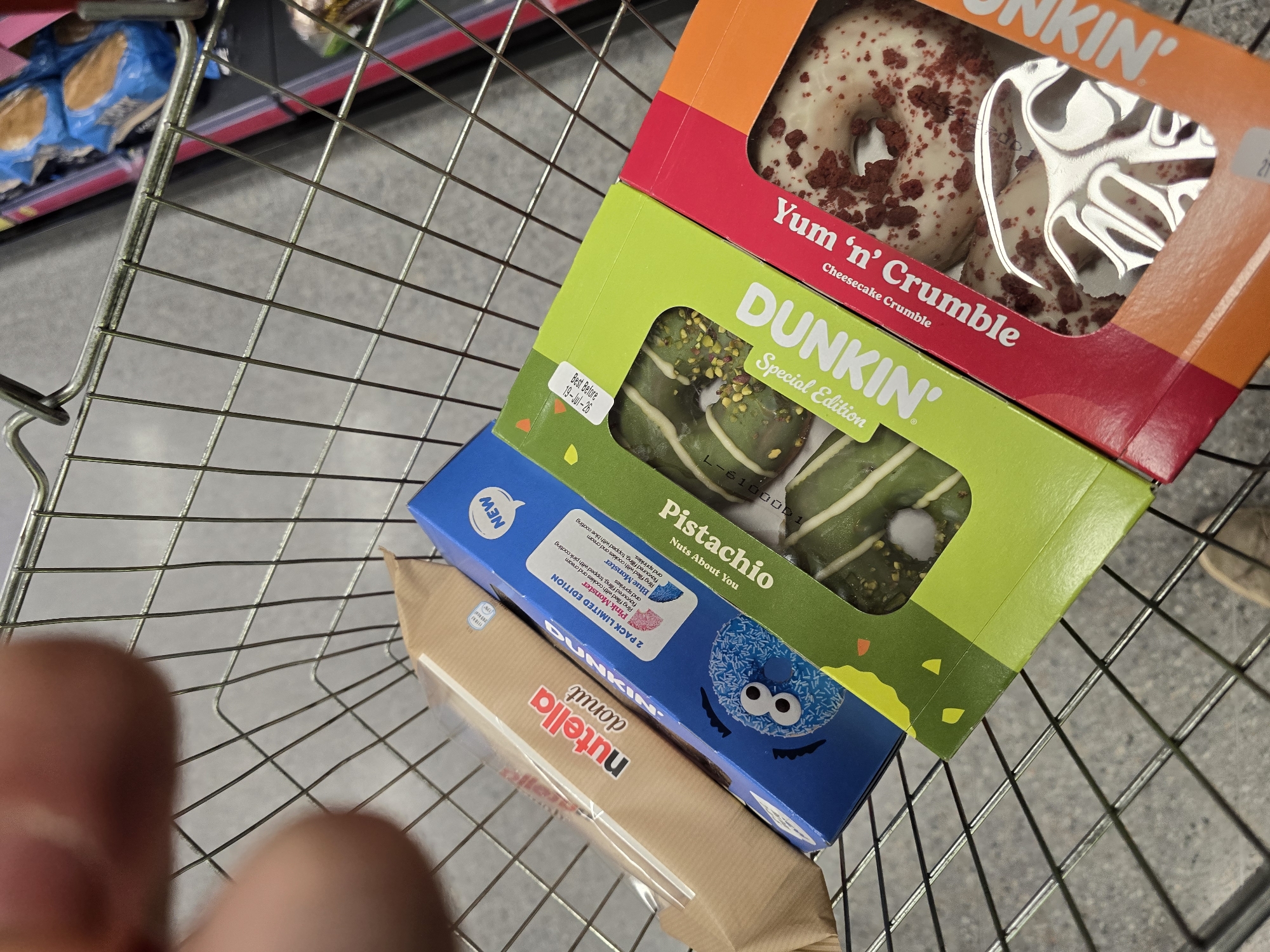 A shopping cart contains a brown paper-wrapped box of Nutella donuts, a blue box of two Dunkin' donuts labeled "Pink Monster" and "Blue Monster," a green box of "Pistachio" donuts, and a red and orange box of "Yum 'n' Crumble" donuts. The donuts are visible through the packaging. The shopping cart is on a tiled floor. The image is taken from above, looking down into the cart.