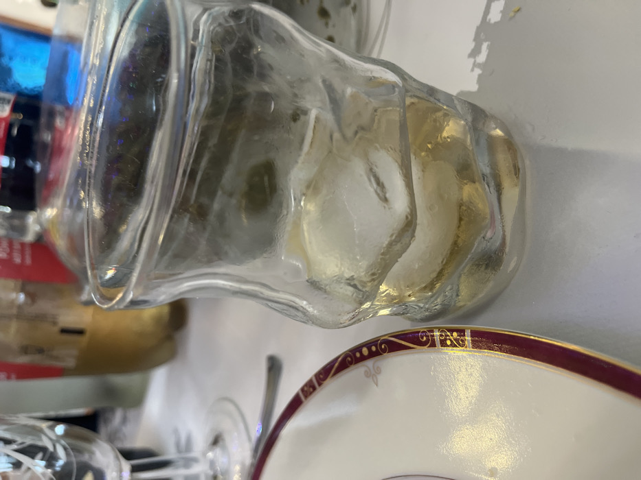 The scene is set on a white tabletop in Ancenis, France. A clear, irregularly shaped glass tumbler occupies the central foreground, containing a pale yellow liquid and approximately three discernible ice cubes. To the immediate left, a portion of a white ceramic plate is visible, featuring a decorative dark red rim adorned with gold scrollwork and dots. A silver-toned utensil handle is positioned between the plate and the tumbler. In the blurred background, several bottles are present: one appears amber-colored, another exhibits a red label, and a third shows a distinct blue hue. A stemmed, clear glass with etched patterns is partially visible on the far left. No individuals or direct actions are depicted.