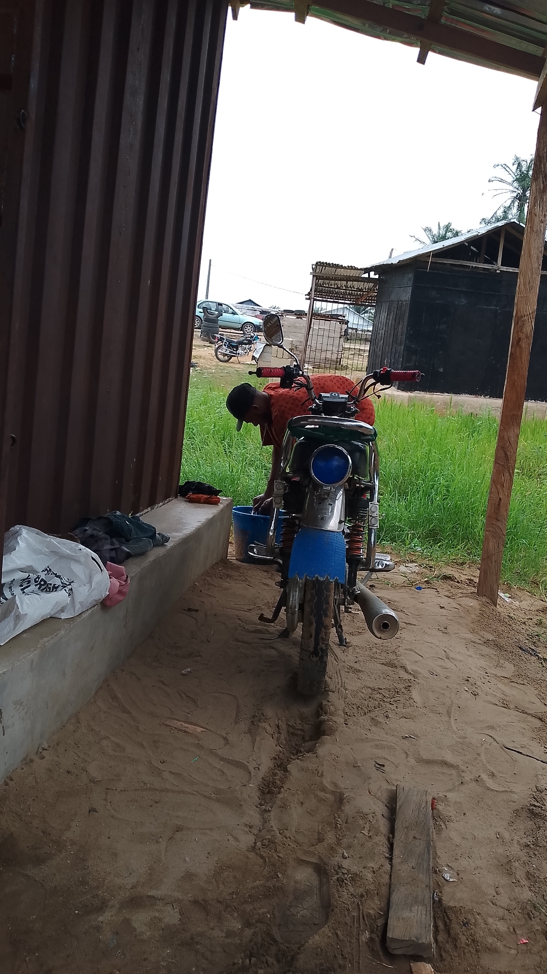 The image depicts a man washing clothes next to a parked motorcycle. The man, wearing a red shirt and a black cap, is bent over a blue bucket of water. The motorcycle, with a green gas tank, chrome accents, and a blue fender flap, is parked on a dirt or sandy surface. Behind the motorcycle is a field of green grass. To the left, there is a building with dark brown corrugated metal siding and a concrete base. The location appears to be in a rural or semi-rural area. In the background, there is another building and a car parked along a road. Based on the prompt, the location is in Asubi, Nigeria.